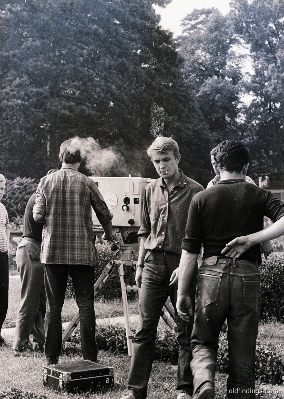 1960s outdoor film production scene: crew operates vintage 16mm camera with tripod in wooded area. Smoking and plaid shirts reflect mid-century filmmaking culture.