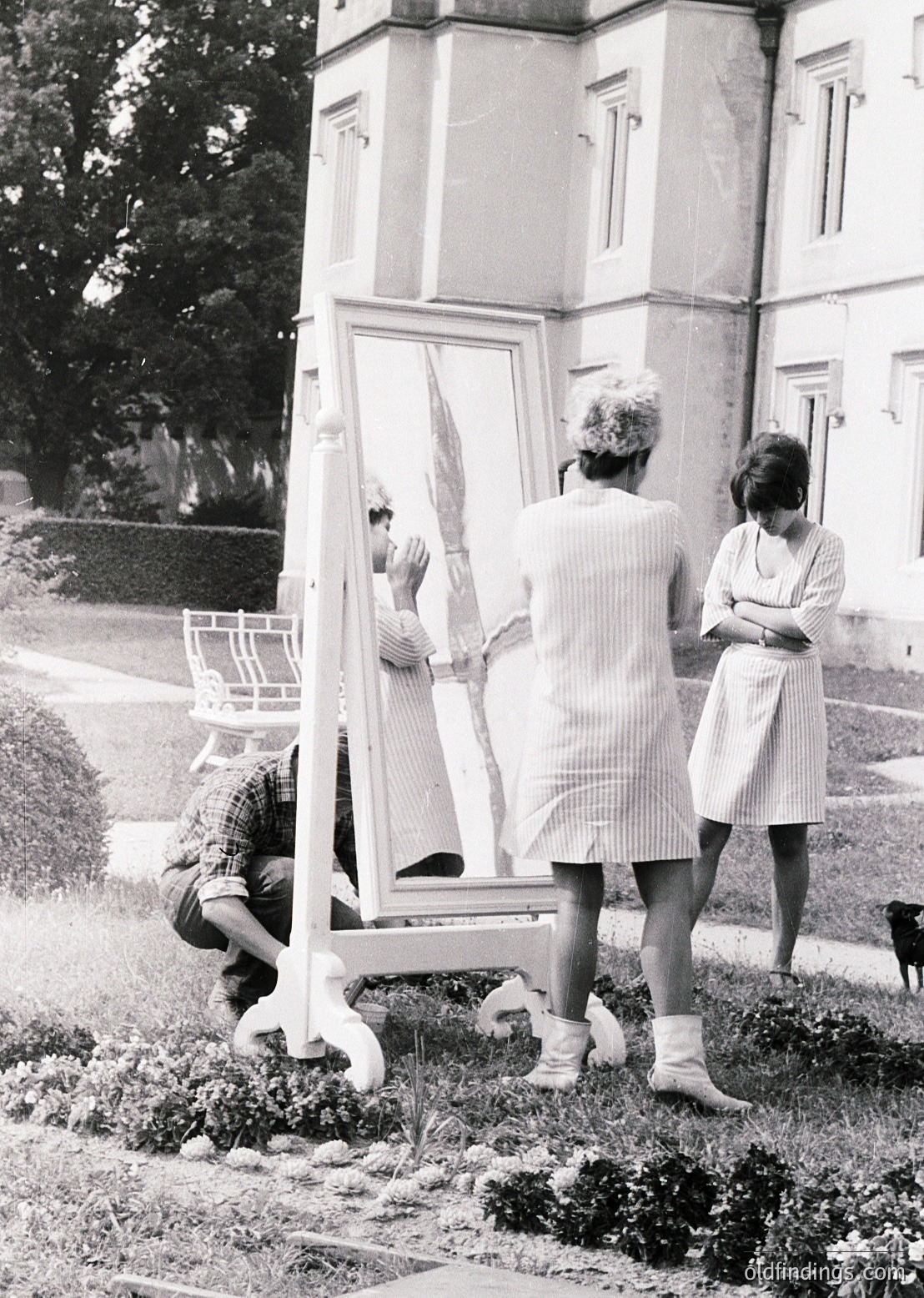 Vintage black-and-white photo of three women in 1960s-era attire posing with a large handheld mirror outdoors. The woman kneeling wears a checkered blouse and white apron, while the others in striped dresses pose near a grand, classical-style building with arched windows. A small dog sits nearby on gravel.