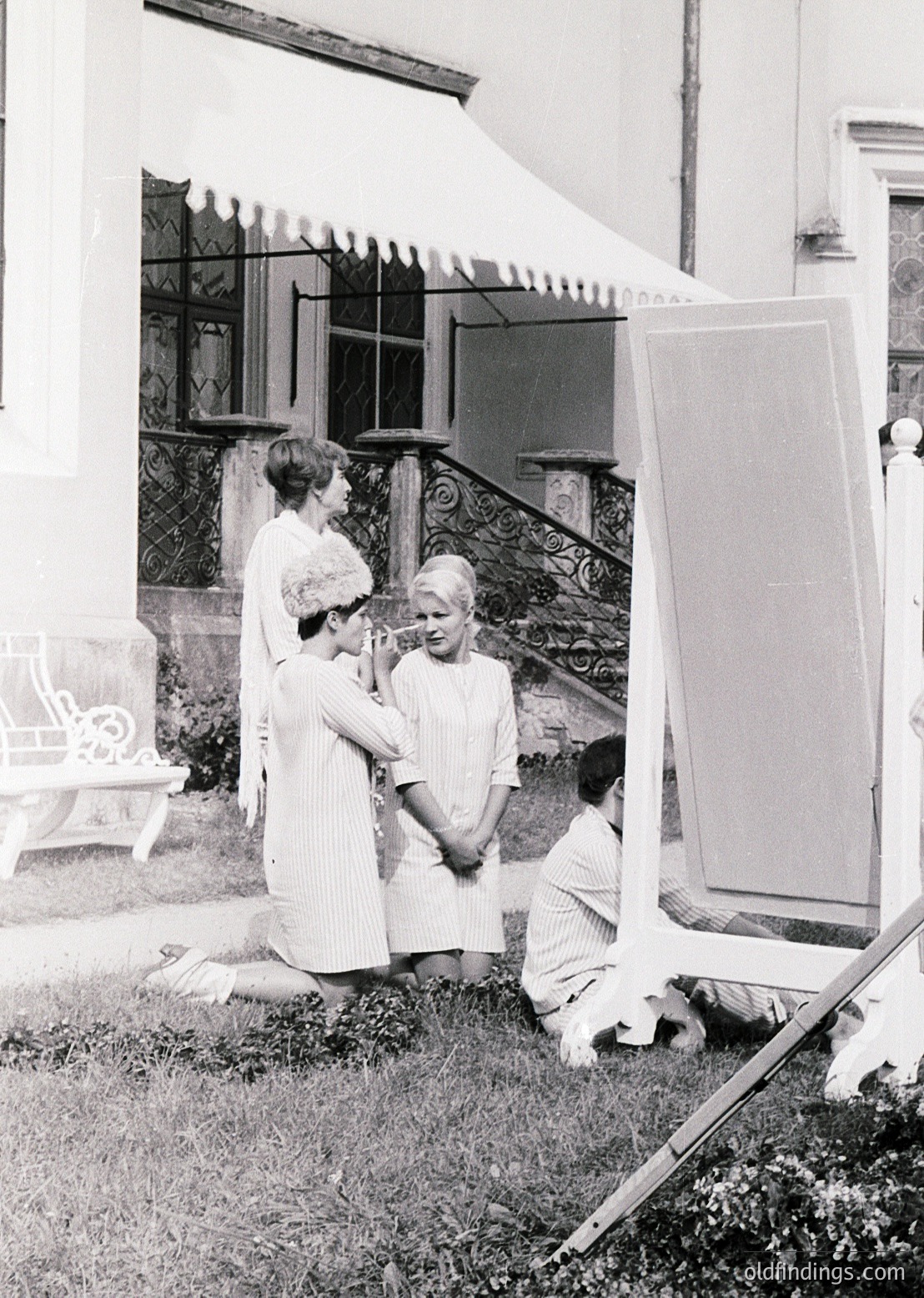 Vintage black-and-white photo of three women in 1960s-era attire—striped dresses, sunglasses, and hairstyles—posing outdoors near ornate wrought-iron railings and a grand staircase. One woman kneels with a small dog, while another adjusts the third’s hair. Reflective mirror and vintage camera equipment suggest a portrait session. Residential architecture hints at mid-century European or American suburban style.