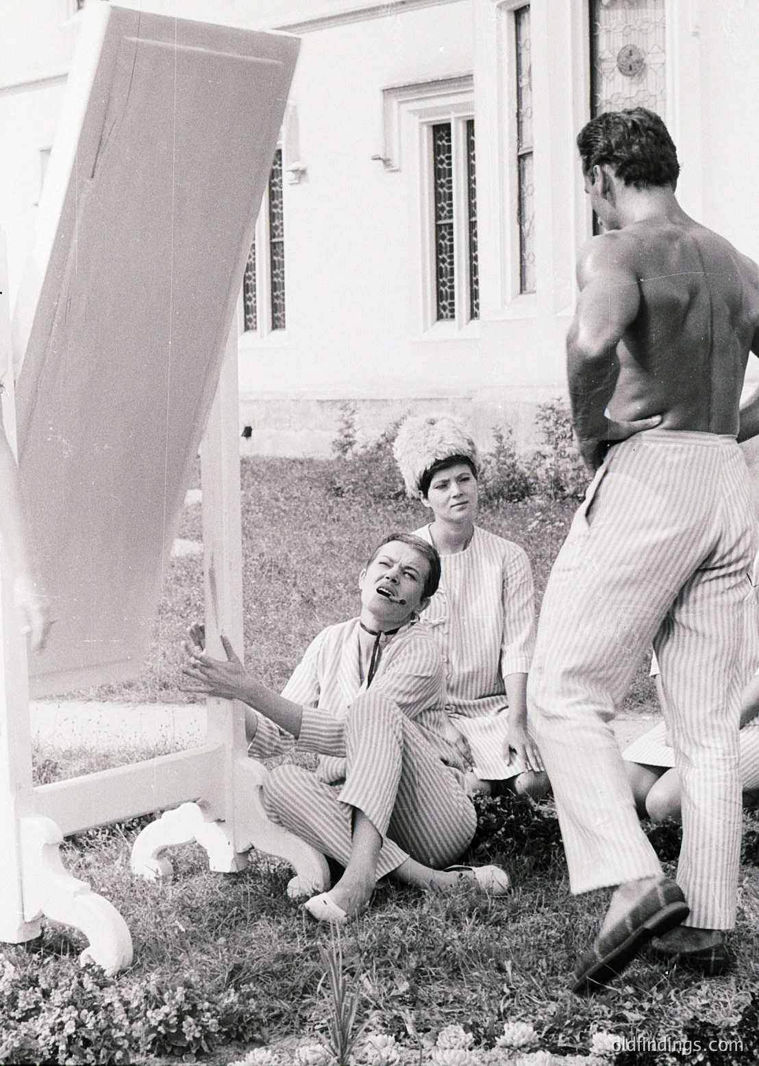 Vintage black-and-white photo of three individuals posing with a large, open, white folding screen in an outdoor setting. The woman kneeling wears a striped dress and a pillbox hat, while the man standing wears loose trousers and no shirt. The background features a building with wrought-iron balconies and lush greenery. Likely mid-20th century, possibly 1950s–1960s.