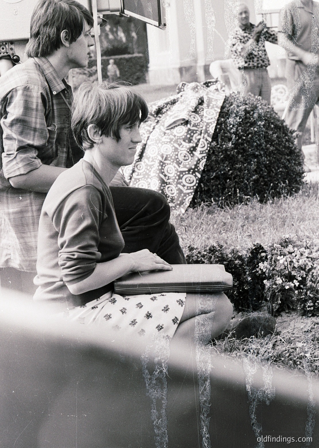 Black-and-white street scene from the 1960s–70s, featuring a young man seated on a low wall, engrossed in a book. He wears a short-sleeve button-up shirt and patterned shorts. Background shows blurred pedestrians, a patterned umbrella, and urban greenery. Ideal for vintage lifestyle, education, or urban history research.