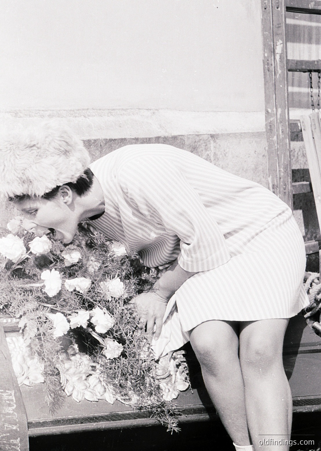 A woman in a 1960s-style striped dress arranges a floral wreath, likely for a memorial or funeral. The setting appears to be an indoor space with a wooden casket and metal railing. The wreath features white flowers and greenery, evoking a solemn, respectful tone.