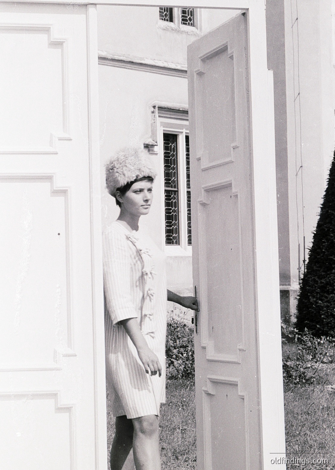 Mid-century woman in a fitted, floral-patterned dress and voluminous bouffant hairstyle poses beside a white door with vertical panels. Classic 1960s fashion and architecture suggest a suburban or residential setting. The image captures timeless elegance and retro aesthetics.