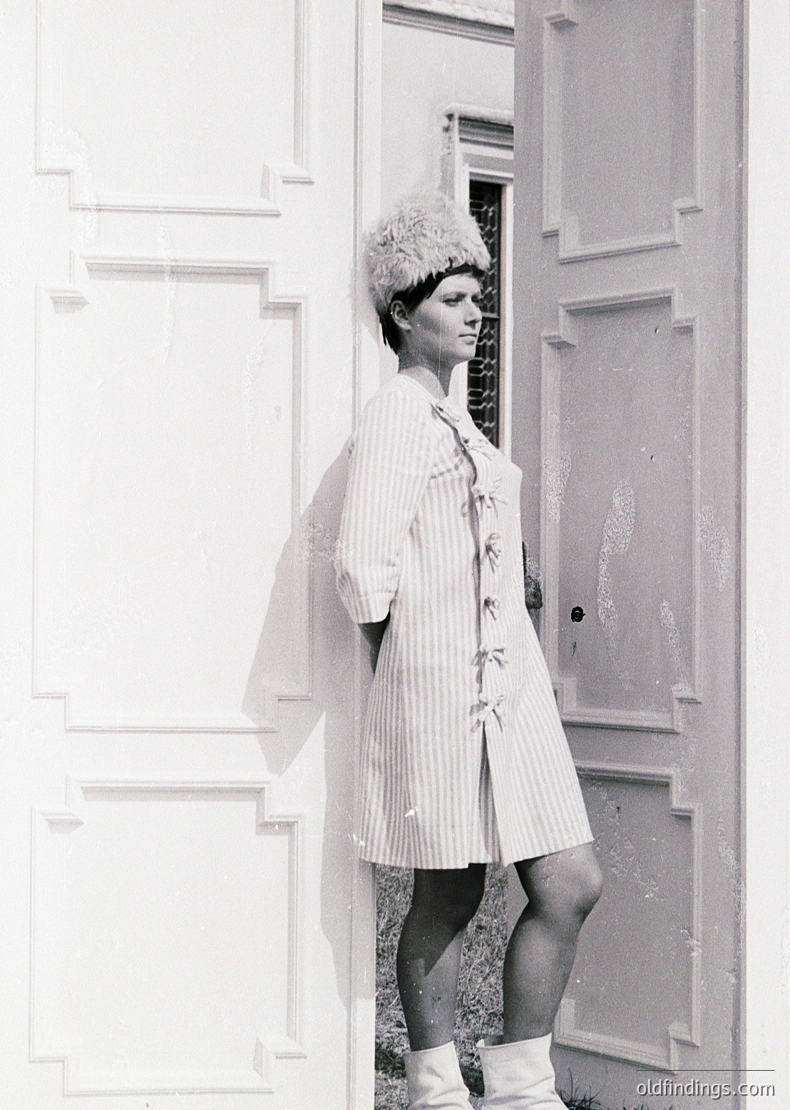 Vintage black-and-white portrait of a woman in a striped housecoat and fur hat, leaning against a paneled door. The architectural details suggest a mid-century European interior, likely 1950s–1960s.