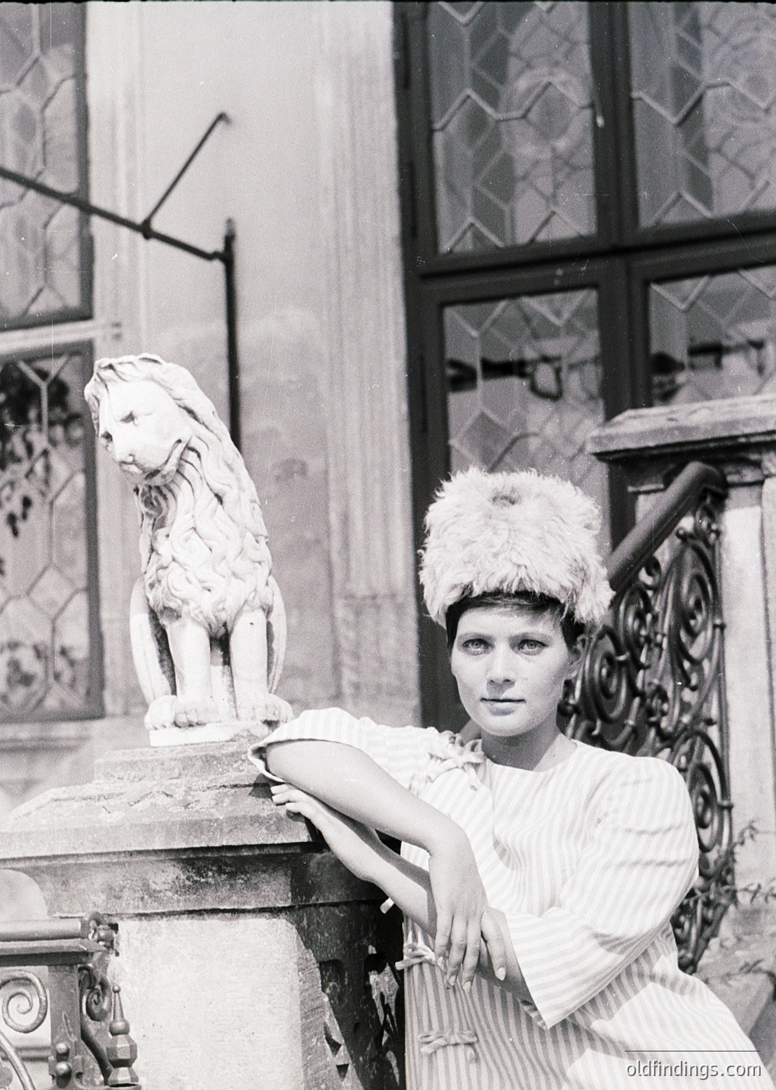 Black-and-white portrait of a woman in 1950s-60s fashion, posing beside a marble lion statue in front of ornate wrought-iron gates. She wears a fur-trimmed headpiece, a striped dress, and a floral brooch. Architectural details suggest a European urban setting.