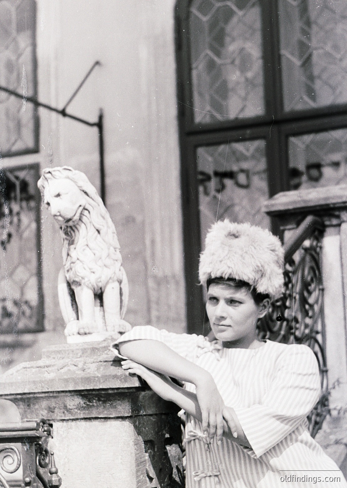 Vintage black-and-white portrait of a woman in 1920s-30s fashion, posing beside a marble lion statue atop an ornate pedestal. She wears a fur-trimmed headpiece and a striped apron over a blouse. Architectural details suggest an urban European setting.