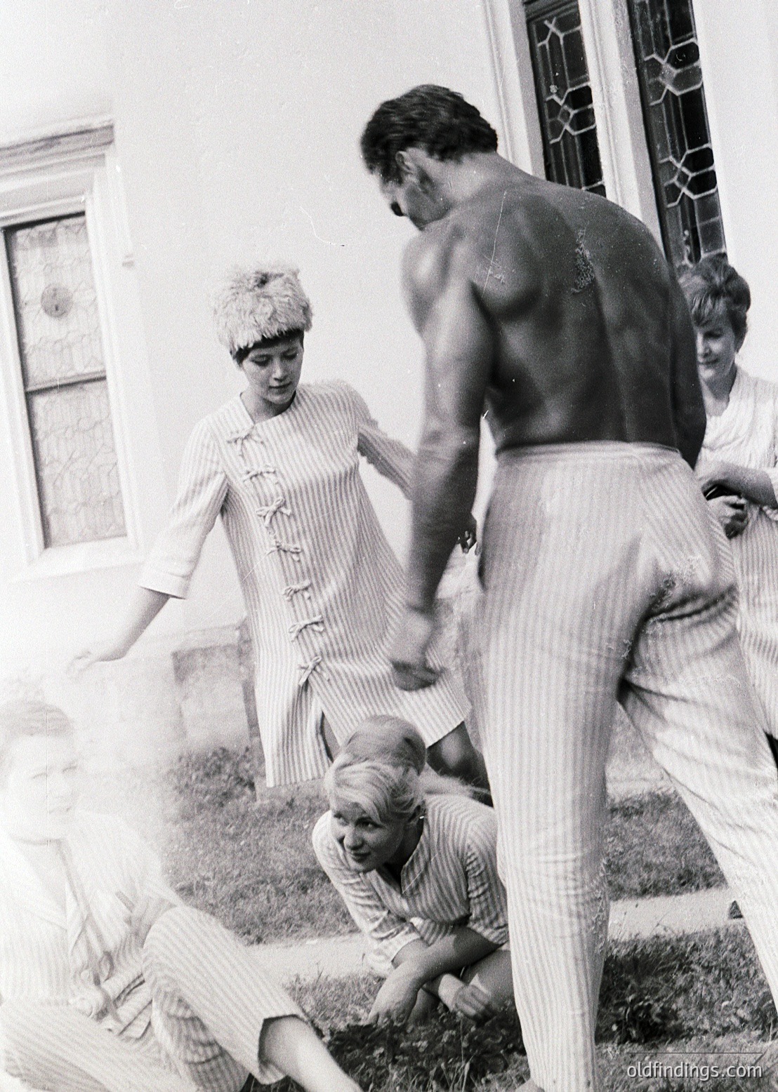 Mid-20th century black-and-white photo capturing a playful moment: a shirtless man in striped pants lifts a child in a striped shirt/shorts, while another child kneels on grass. A woman in a striped blouse with a belted waist stands nearby. Classic 1950s-60s family recreation in a residential backyard.