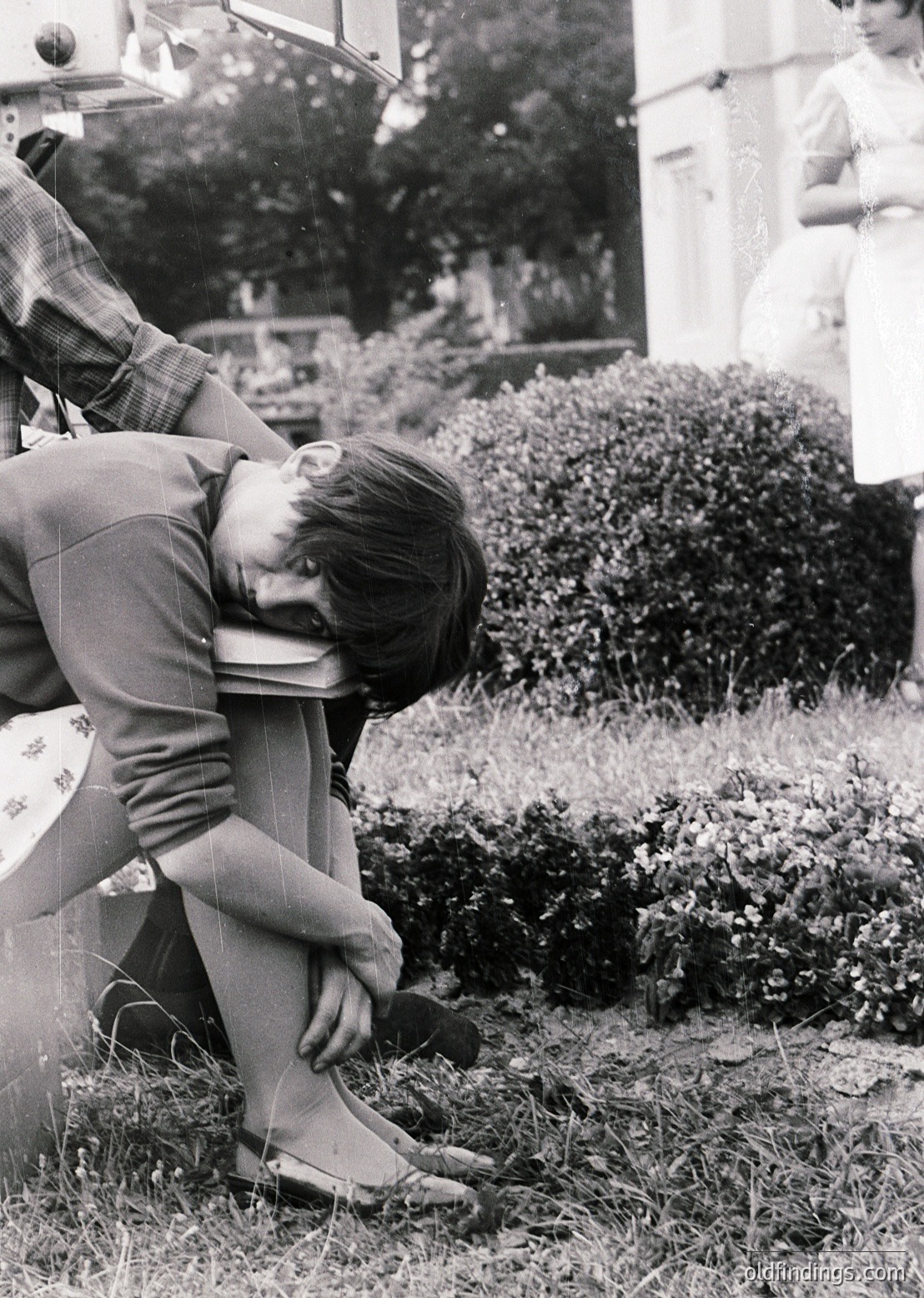 Black-and-white candid of a seated man in 1960s-era clothing (plaid shirt, rolled trousers) adjusting his tie outdoors. Lush greenery and a blurred figure in the background suggest a garden or park setting. Reflective surface (likely a mirror or camera) captures his reflection, adding depth. Ideal for vintage fashion, candid photography, or historical research.