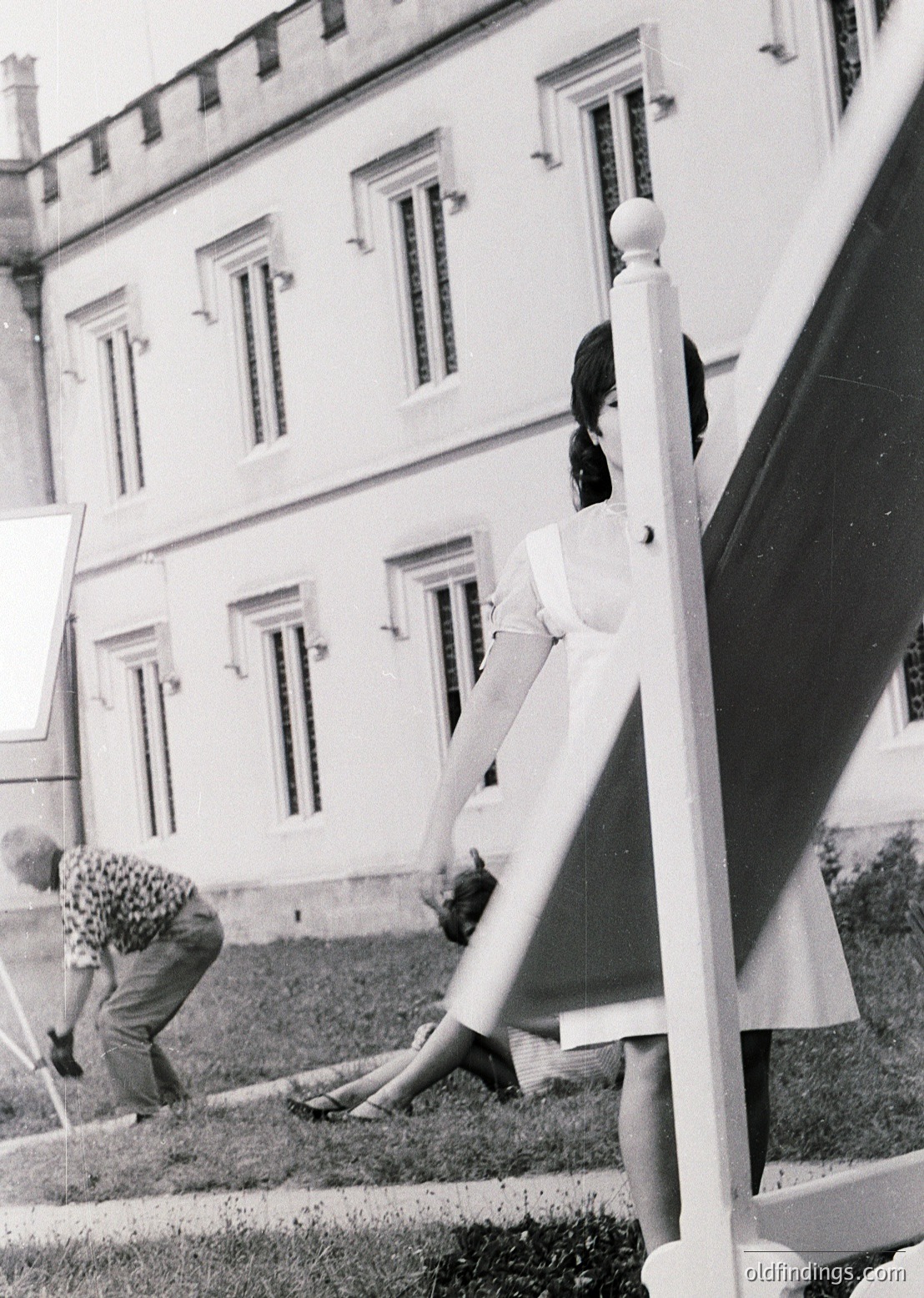 Neatly cropped black-and-white photo of two women in 1950s-60s European courtyard. One woman in a fitted dress and heels leans on a white railing, while another in a floral hat and skirt kneels gardening. Symmetrical building with rectangular windows and decorative moldings in background.