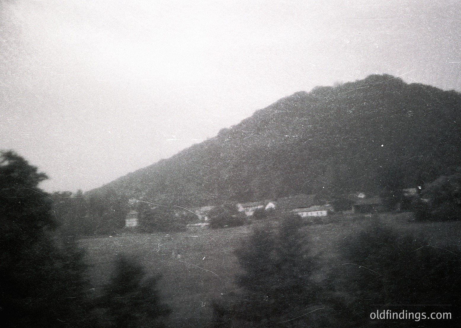 Vintage black-and-white rural landscape featuring modest wooden houses nestled at the base of a forested hillside. Fog or mist obscures mid-ground details, enhancing the serene, timeless atmosphere. Likely early-to-mid 20th century, possibly Eastern Europe.