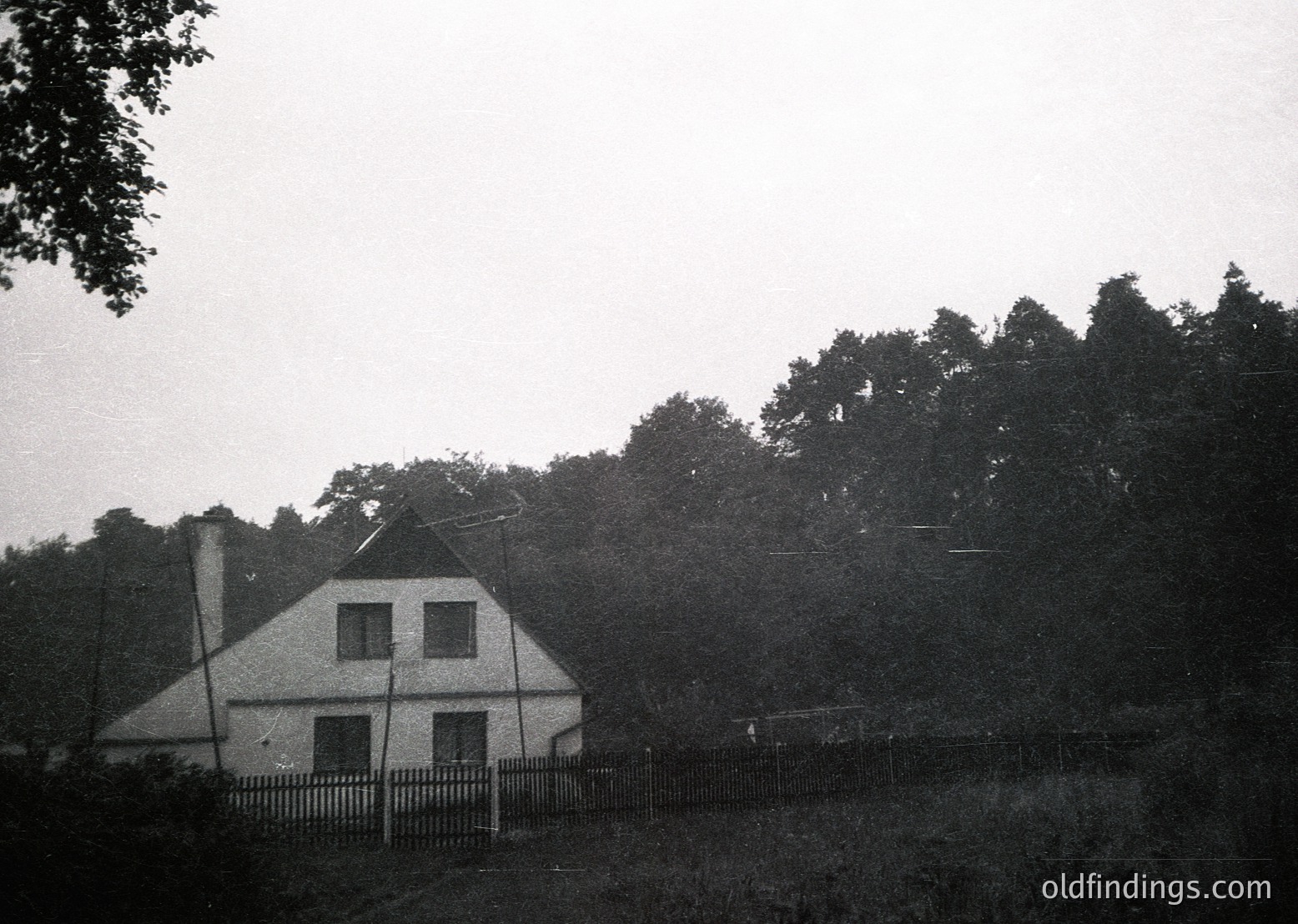 Vintage black-and-white photo of a modest, two-story farmhouse with a gabled roof and chimney, surrounded by dense forest. Fenced yard with overgrown grass suggests rural isolation. Likely mid-20th century European countryside.