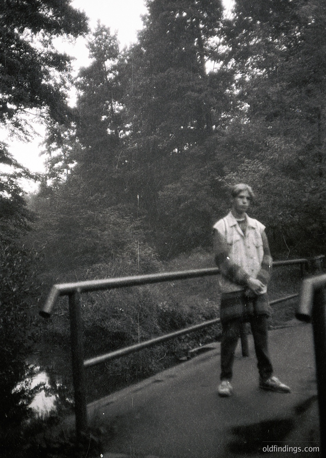 Black-and-white portrait of a young man standing on a metal railing bridge, surrounded by dense forest foliage. His jacket, trousers, and shoes suggest mid-20th-century style. Likely taken in a European park or hiking trail during the 1950s–1970s.