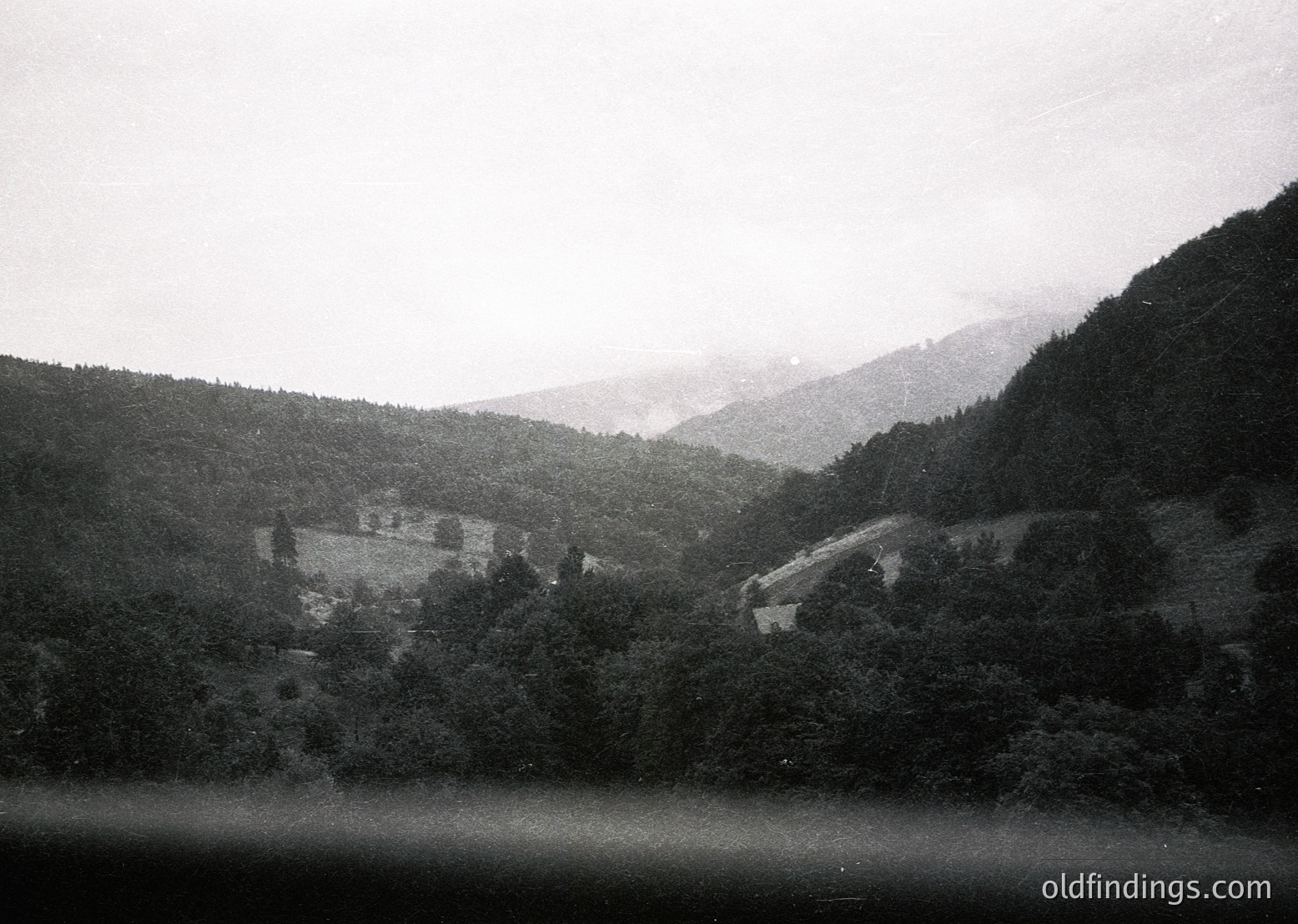 Vintage black-and-white mountain village nestled between dense forests and rolling hills, likely European alpine region. Stone buildings clustered near a winding road, framed by misty peaks. Early 20th-century rural architecture with dense greenery. Ideal for historical research or alpine-themed design.