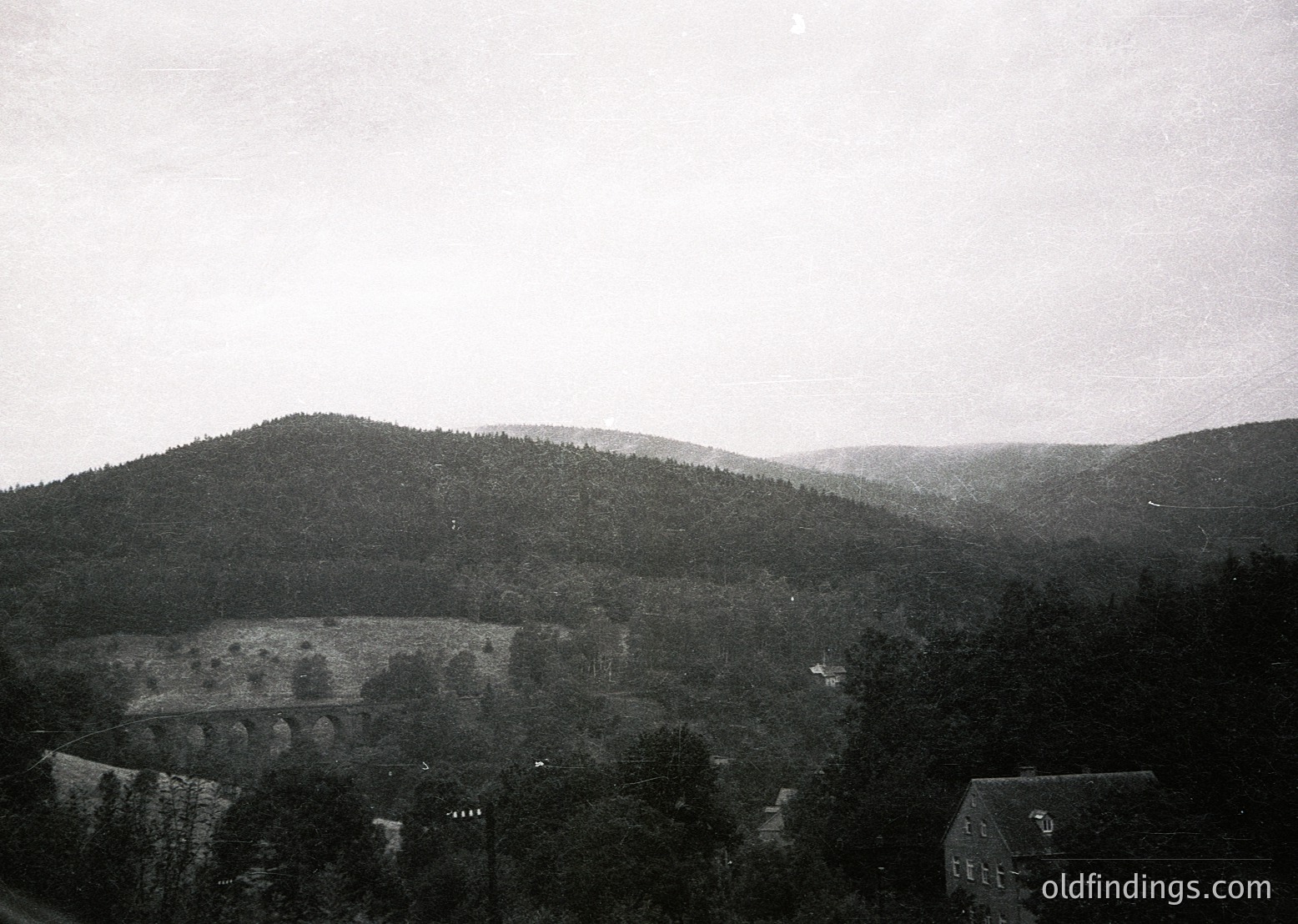 Vintage black-and-white landscape featuring a stone bridge spanning a valley, flanked by dense forest and rolling hills. Mid-20th century architectural elements visible in foreground buildings, likely residential or institutional. Misty atmosphere suggests early morning or overcast conditions. Ideal for historical or nostalgic stock imagery.