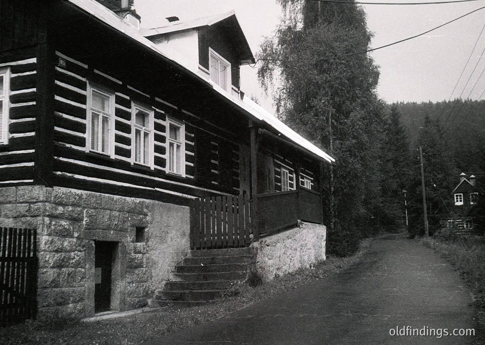 Two-story timber-framed house with stone foundation, set on a rural road. Horizontal wooden beams and white-framed windows contrast with dark wood. Stone steps lead to an arched basement entrance. Dense forest and another house visible in background. Likely Eastern European rural architecture, mid-20th century.