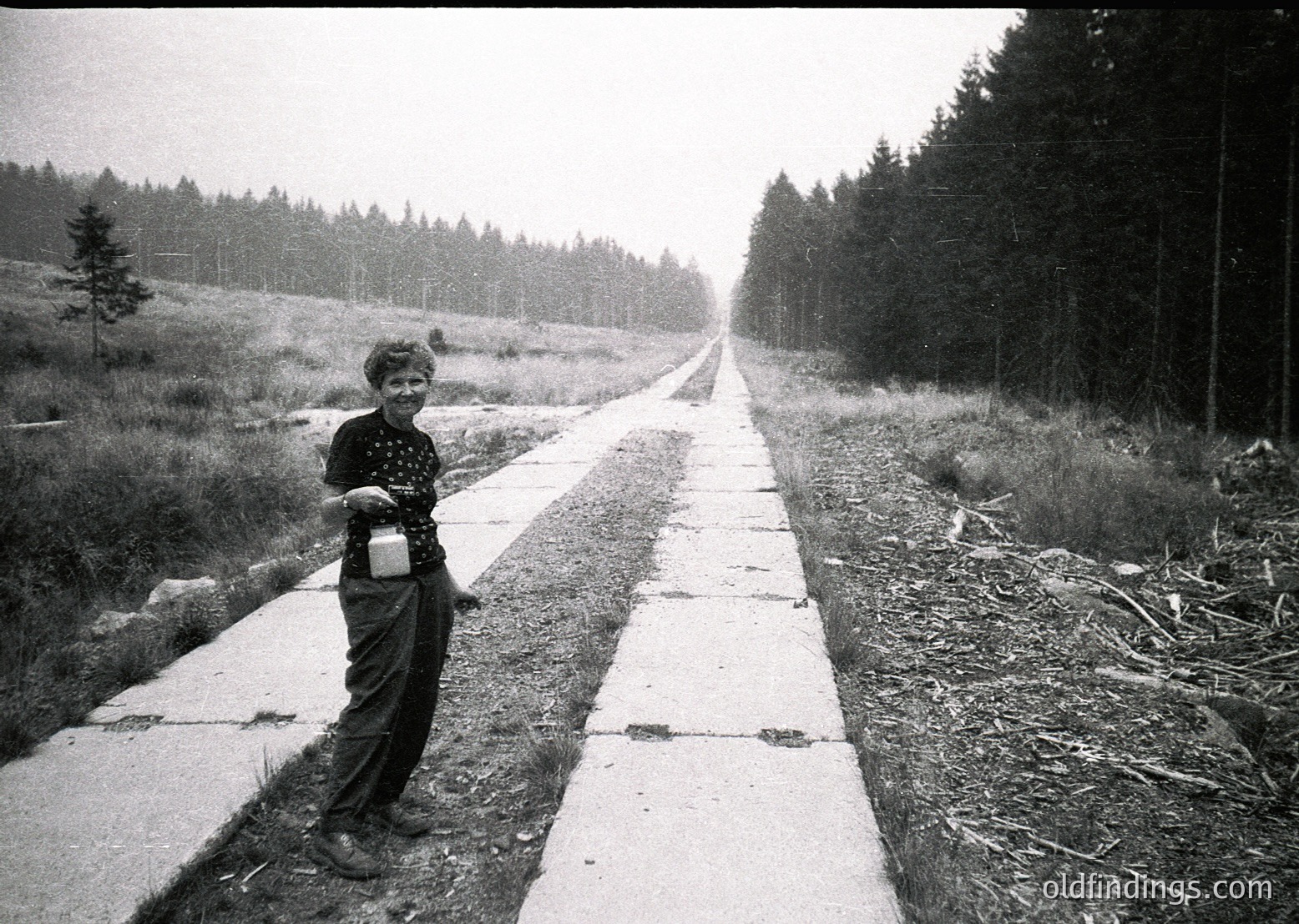 Child in 1960s-70s outdoor setting, holding a small container, standing on a paved path flanked by dense forest. Mid-century concrete sidewalk with visible cracks, leading into misty woodland.