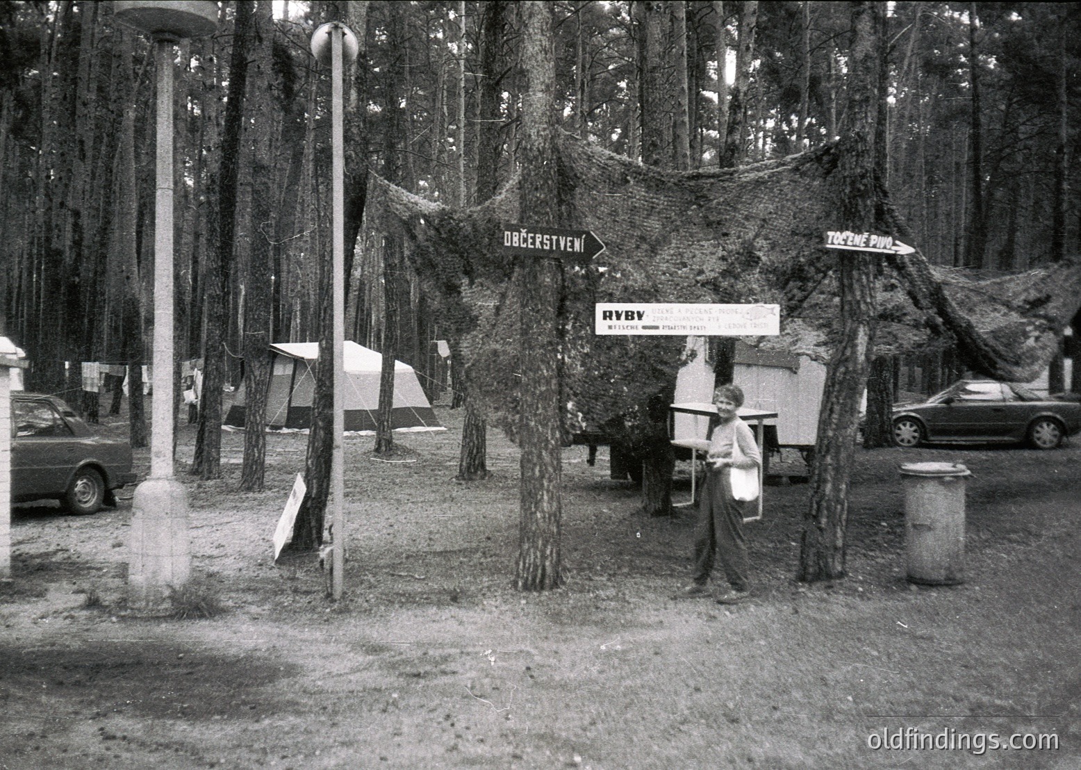 Vintage black-and-white photo of a public park entrance with directional signs in Cyrillic script, likely Bulgaria. A woman in a long dress stands near a small kiosk with a sign reading "РЫБА" (fish). Dense forest and parked cars suggest a mid-20th-century recreational area.