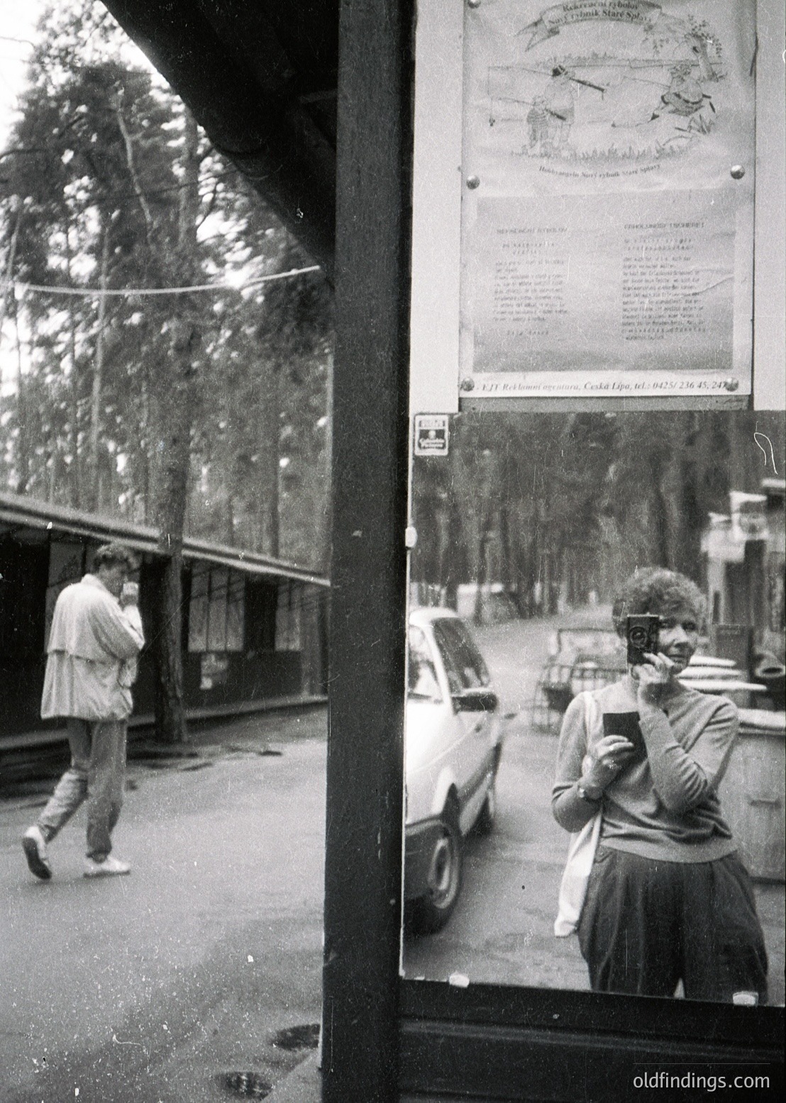 Reflection of a 1970s-era street scene in a shop window: a man in a light jacket walks past parked cars, while a woman in a striped sweater holds a camera. Posters and maps adorn the window frame, suggesting a European urban setting.