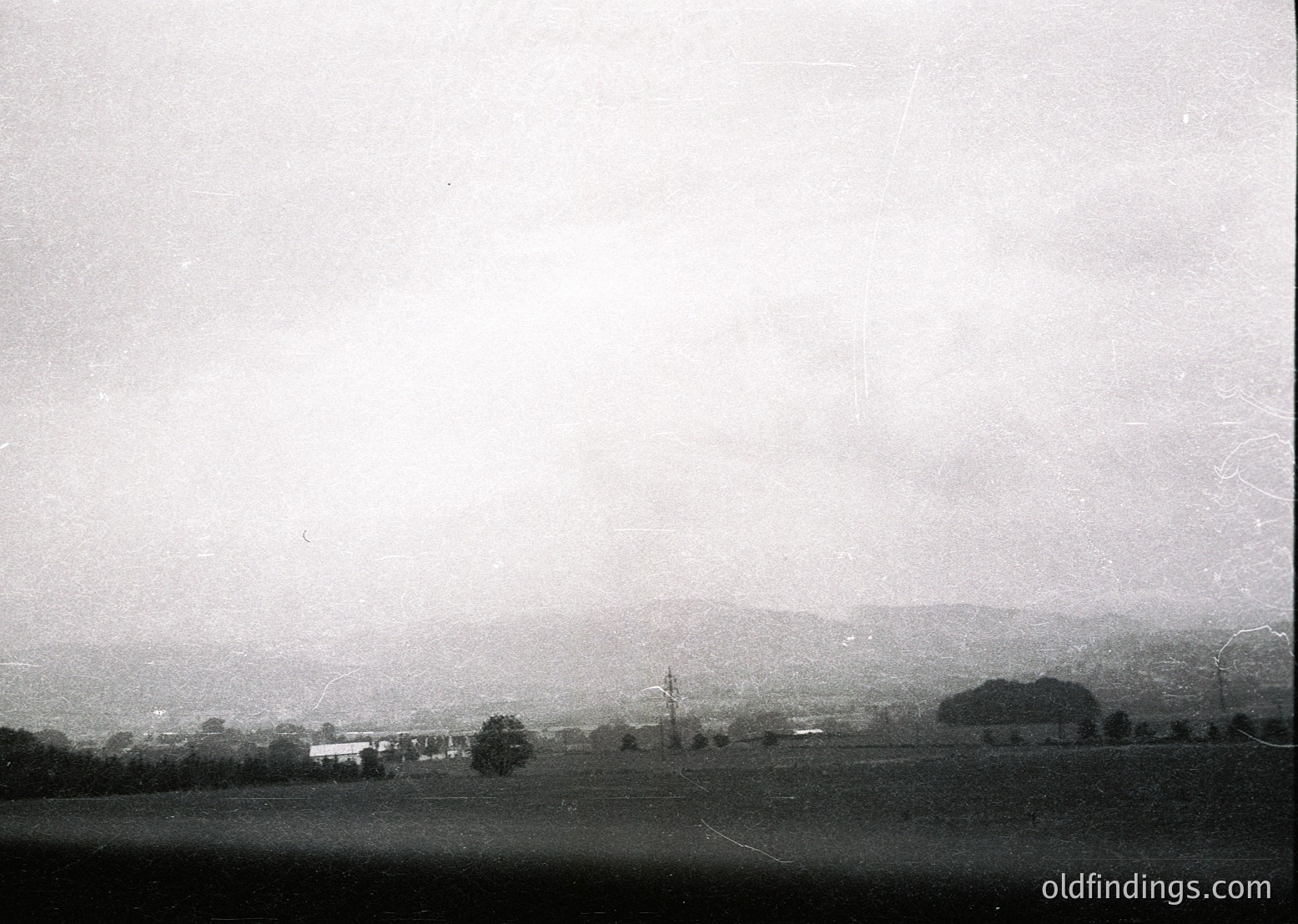 Vintage black-and-white rural landscape featuring sparse farmhouses, scattered trees, and rolling hills under overcast skies. Likely early-to-mid 20th century agricultural setting.