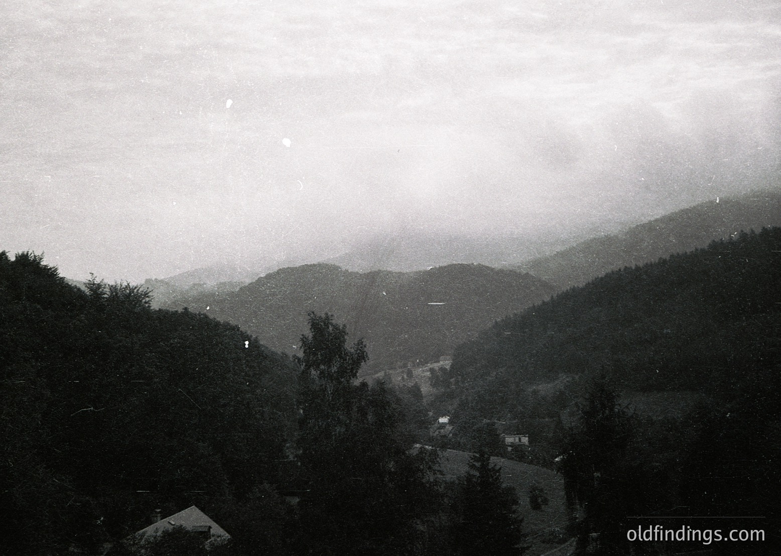 Black-and-white aerial view of a mist-shrouded valley with dense forest and scattered buildings, likely European alpine region. Low-contrast vintage photography suggests early-to-mid 20th century. Ideal for historical research or nostalgic design references.