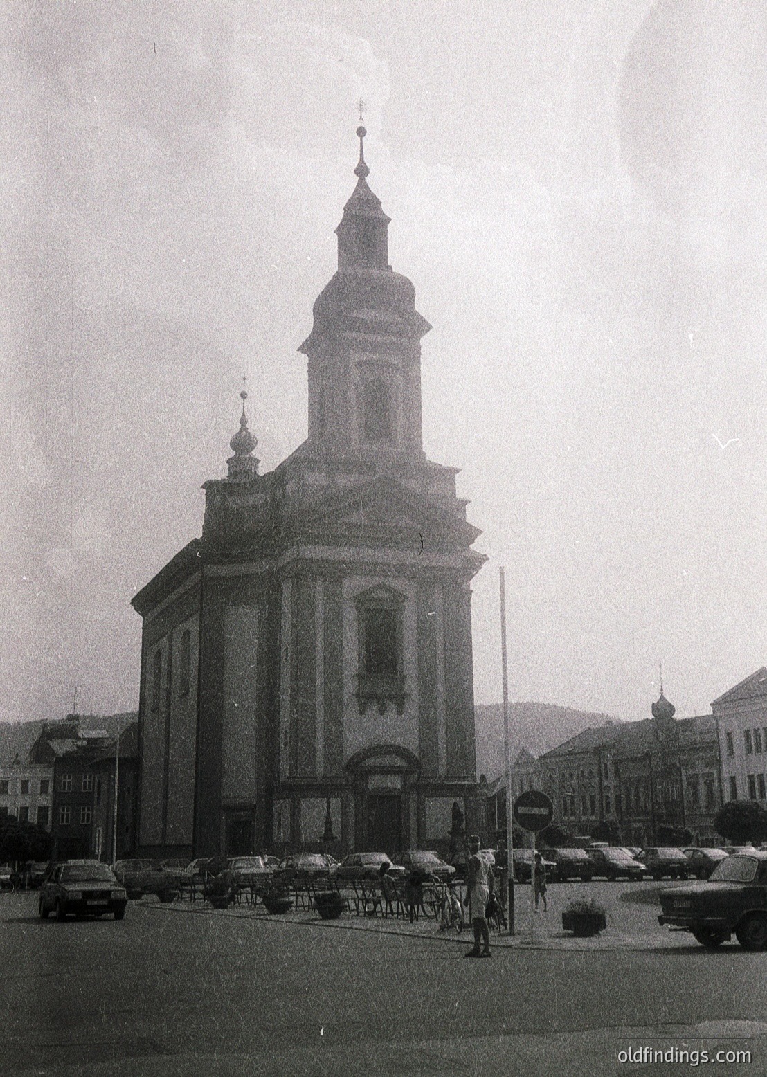 Neoclassical church with twin towers and central spire, likely Eastern European. Symmetrical facade with arched windows and a prominent entrance. Mid-20th century urban setting, featuring vintage cars and minimal pedestrians. Foggy or misty atmosphere enhances architectural details.
