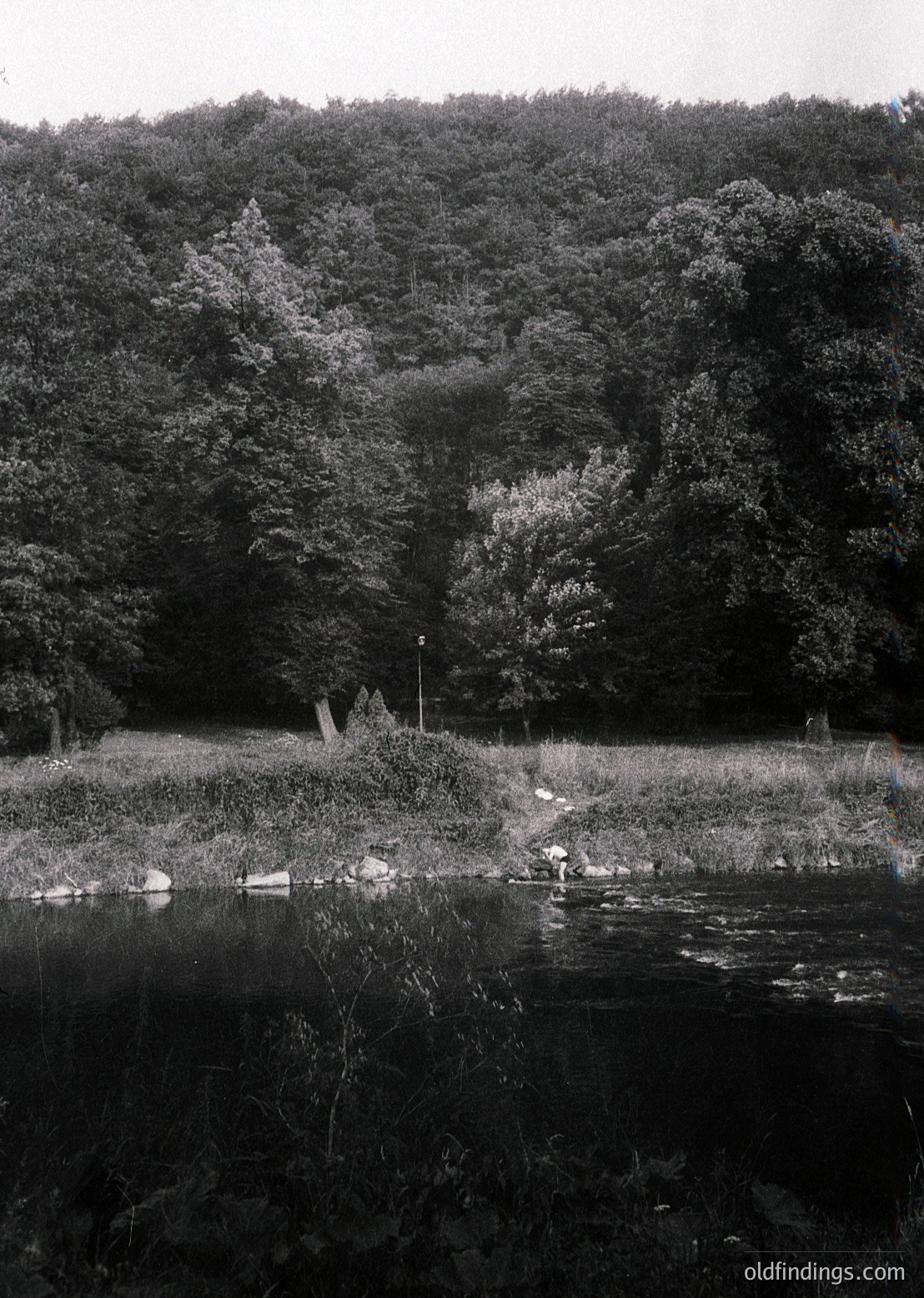 Black-and-white landscape featuring a lone figure standing near a rocky riverbank, surrounded by dense forest. The scene evokes mid-20th century solitude, likely or . Reflections in the water enhance the serene, contemplative mood.