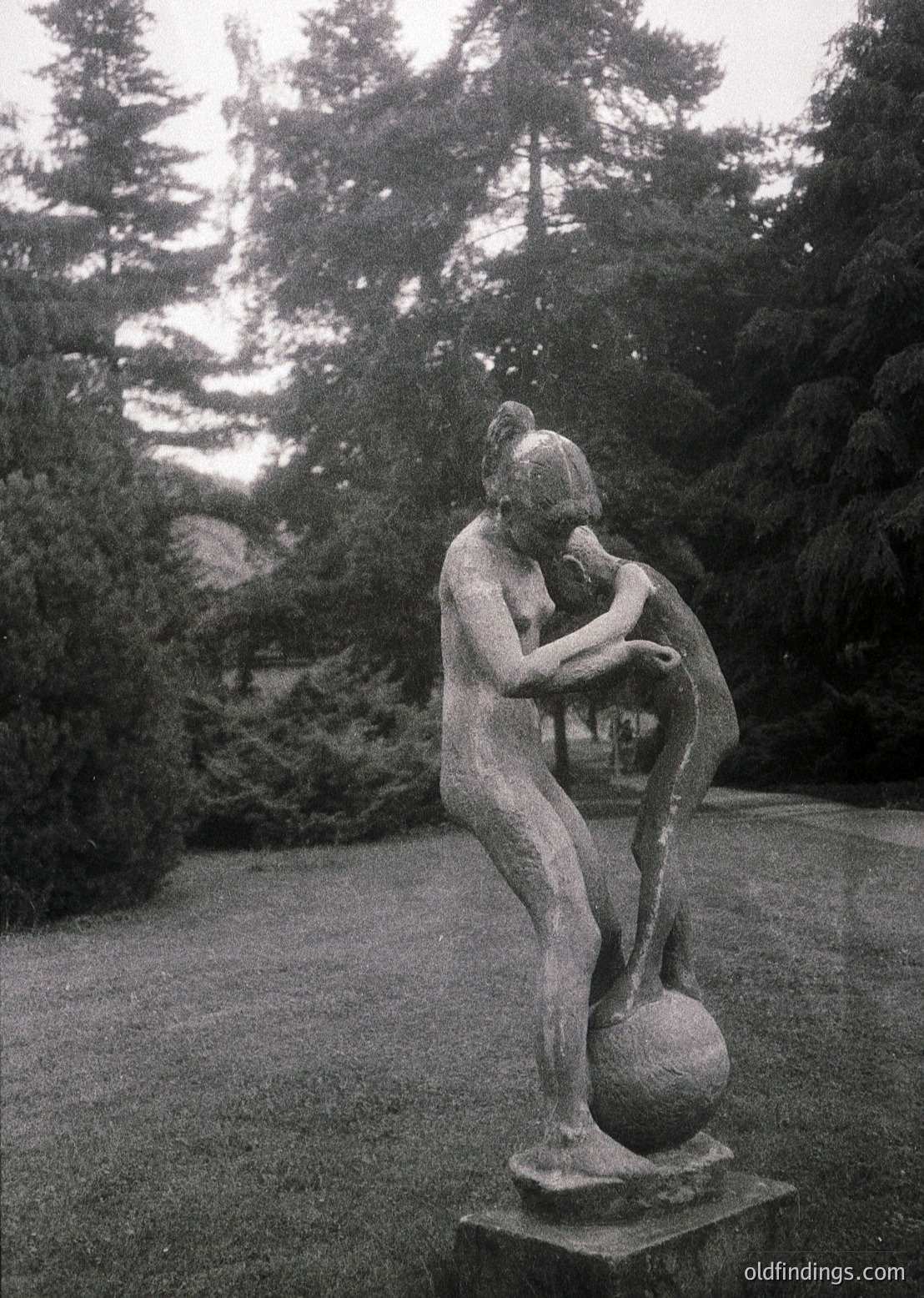 Classic bronze sculpture of a seated woman cradling a child, set in a serene garden. The composition evokes maternal tenderness and timelessness. Likely mid-20th century, European park or estate.