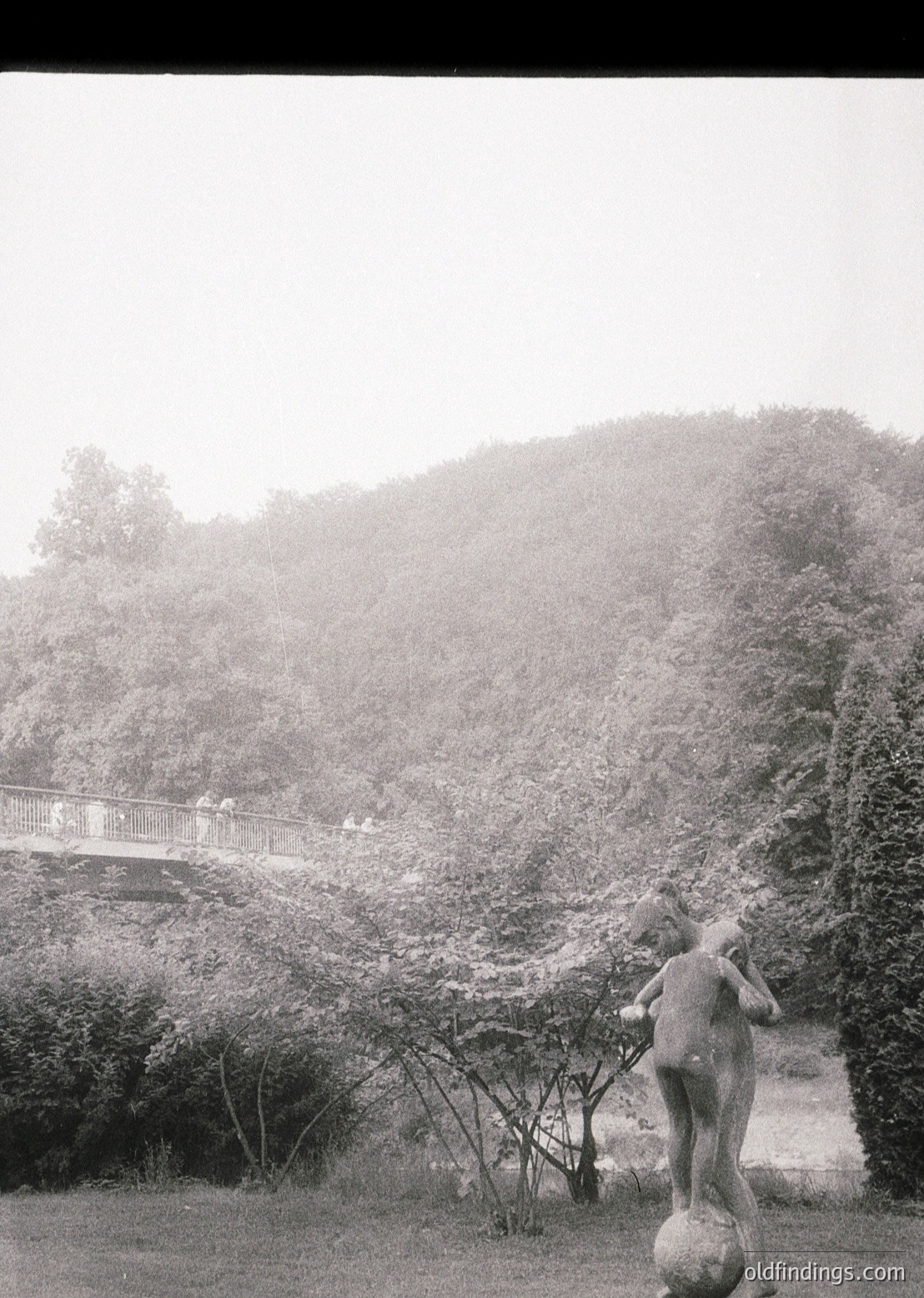 Vintage black-and-white photo of a stone statue depicting a child pushing a large boulder, likely from a public park or garden. Bridge with railings and people in the background atop a forested hill. Foggy atmosphere enhances the nostalgic, mid-20th-century feel.