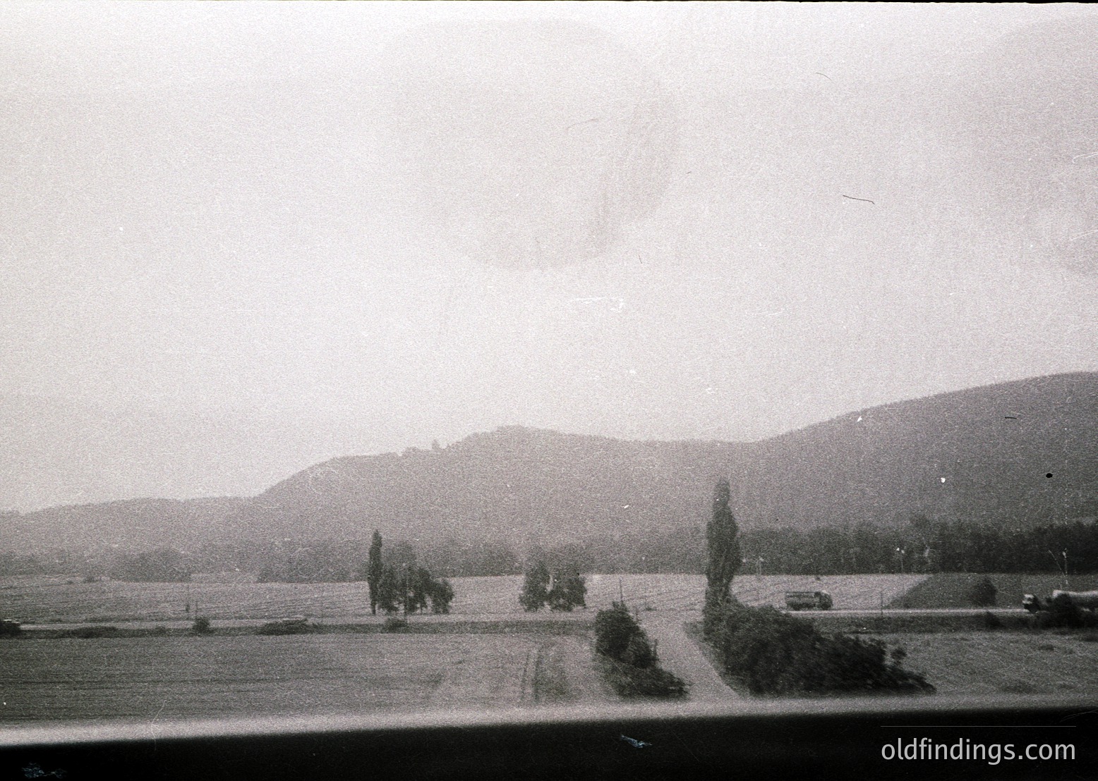 Vintage black-and-white landscape featuring rolling hills, sparse trees, and a lone road winding through open fields. Overcast sky suggests mid-20th century rural setting. Low-angle perspective hints at a window or barrier framing the view.