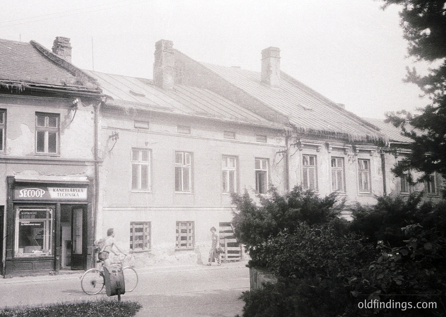 Two-story Soviet-era brick building with symmetrical white-framed windows, likely a commercial/residential hybrid. Signage reads "SLOOP" (shop) and "KANCHERI TEHNIKA" (technical supplies). A man rides a bicycle past, another stands near a wooden fence. Overgrown greenery frames the right side.
