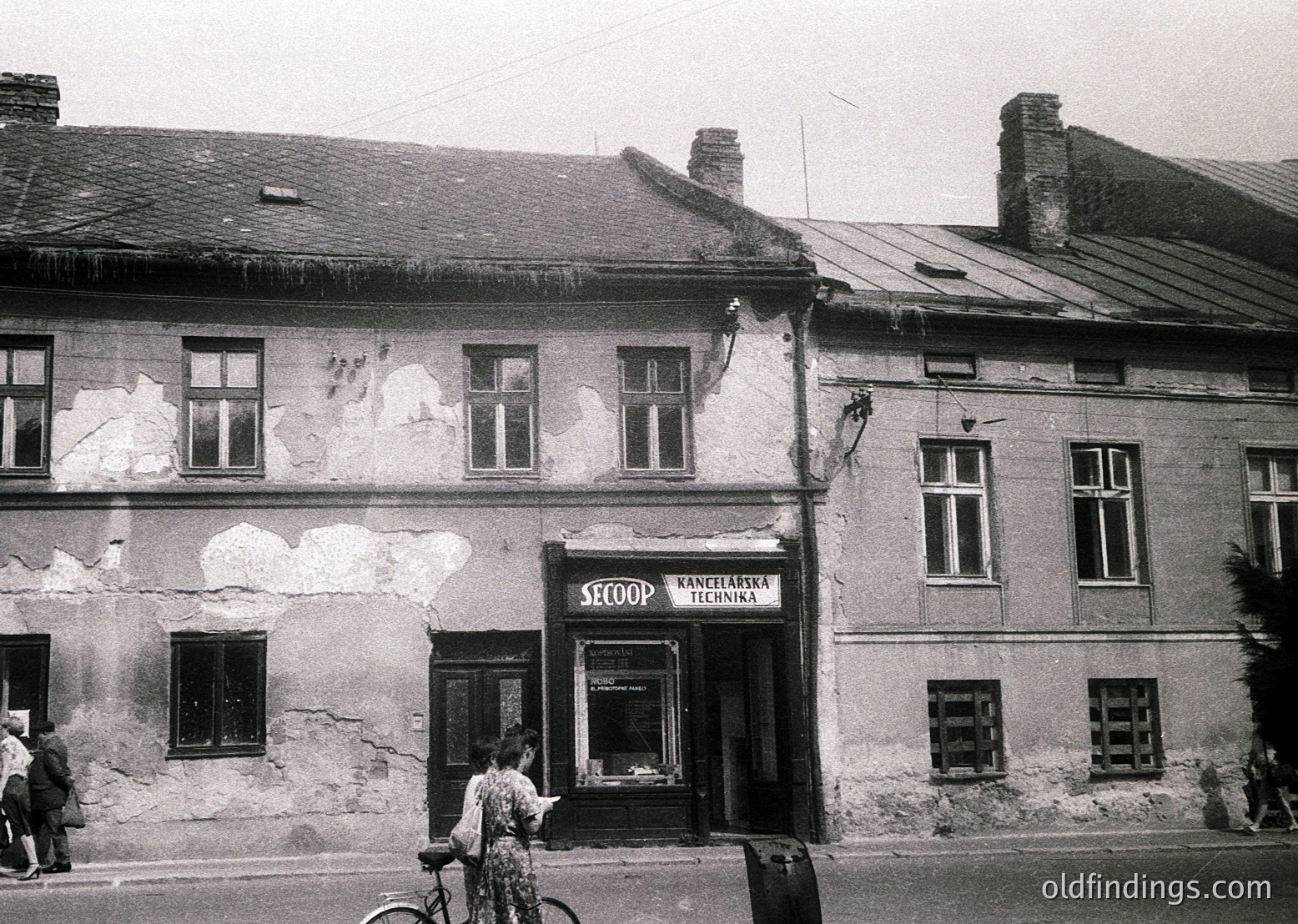 Mid-20th century urban storefront in Eastern Europe, likely Bulgaria. The building’s worn plaster and signage ("SECOOP" + "Разделка Техника") suggest a cooperative-era shop, possibly selling agricultural tools or household goods. Pedestrians and a cyclist add street life context.