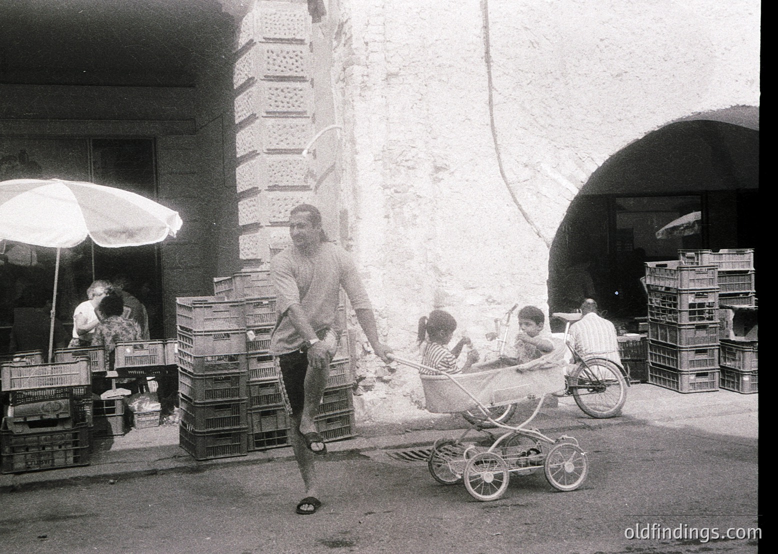 Vintage black-and-white street scene featuring a man pushing a baby stroller while balancing a crate on his shoulder. Stacked crates and a woman seated under an umbrella suggest a bustling market or delivery area. Mid-20th century urban lifestyle captured.