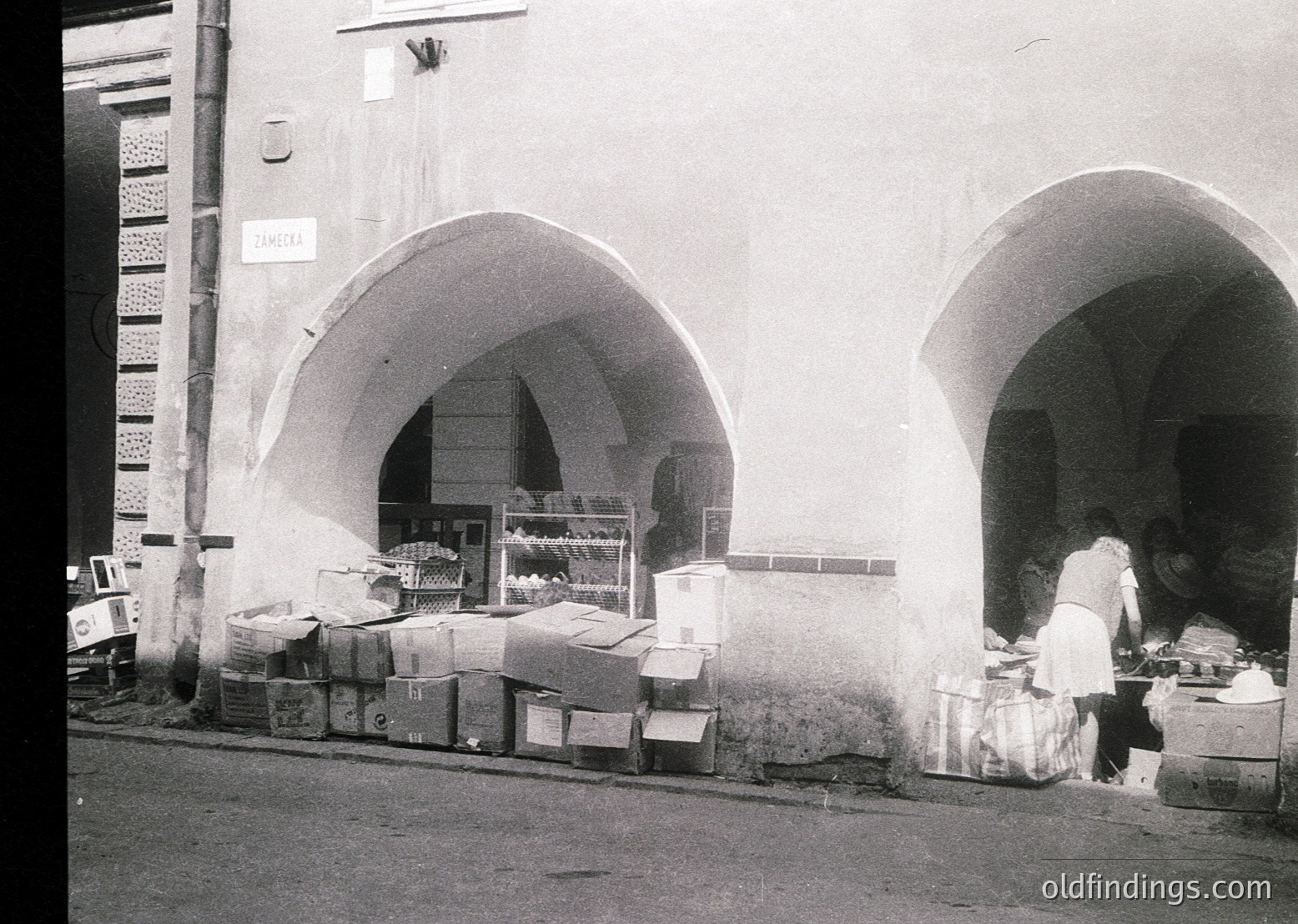 Vintage black-and-white street market scene featuring two arched storefronts with stacked crates and baskets of goods. A vendor in a white apron organizes produce inside the right arch. Decorative tilework on the left wall adds architectural detail. Likely Mediterranean or Middle Eastern, 1950s–1960s.