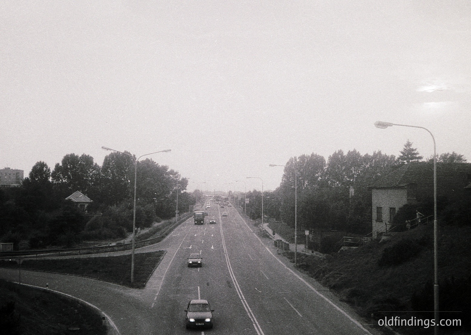 Mid-20th century urban roadway with sparse traffic, flanked by dense greenery and streetlights. Concrete barriers and a single-lane bridge curve into the distance. Overcast skies and monochrome filter evoke a nostalgic, mid-century atmosphere.