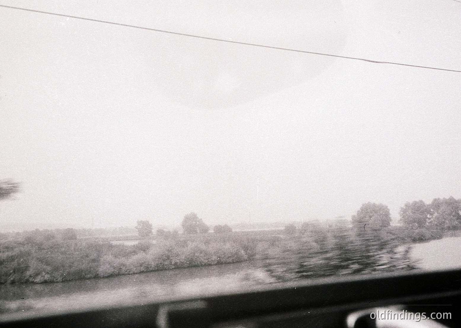 Blurred monochrome shot from a moving vehicle, showing a wet roadside and distant treeline under overcast skies. Raindrops distort the windshield. Likely mid-20th century due to film grain and composition. Ideal for vintage travel or atmospheric mood references.