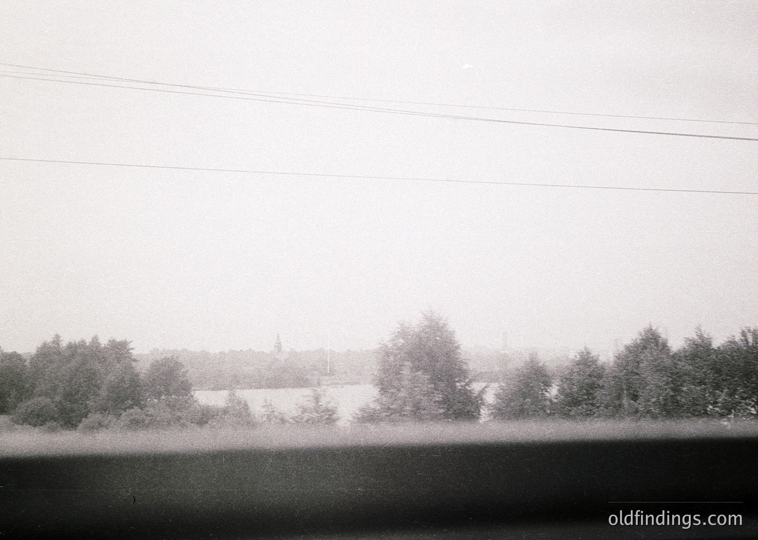 Vintage black-and-white landscape featuring misty horizon with dense tree line and distant church spire. Overcast sky and power lines frame composition. Likely rural European setting, 20th-century style.