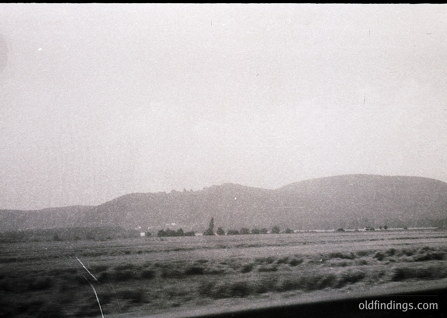 Vintage black-and-white landscape showing rolling hills and sparse vegetation, likely mid-20th century. Flat terrain dominates foreground with a distant village or farm cluster near base of hills. Overcast sky enhances moody, timeless atmosphere.