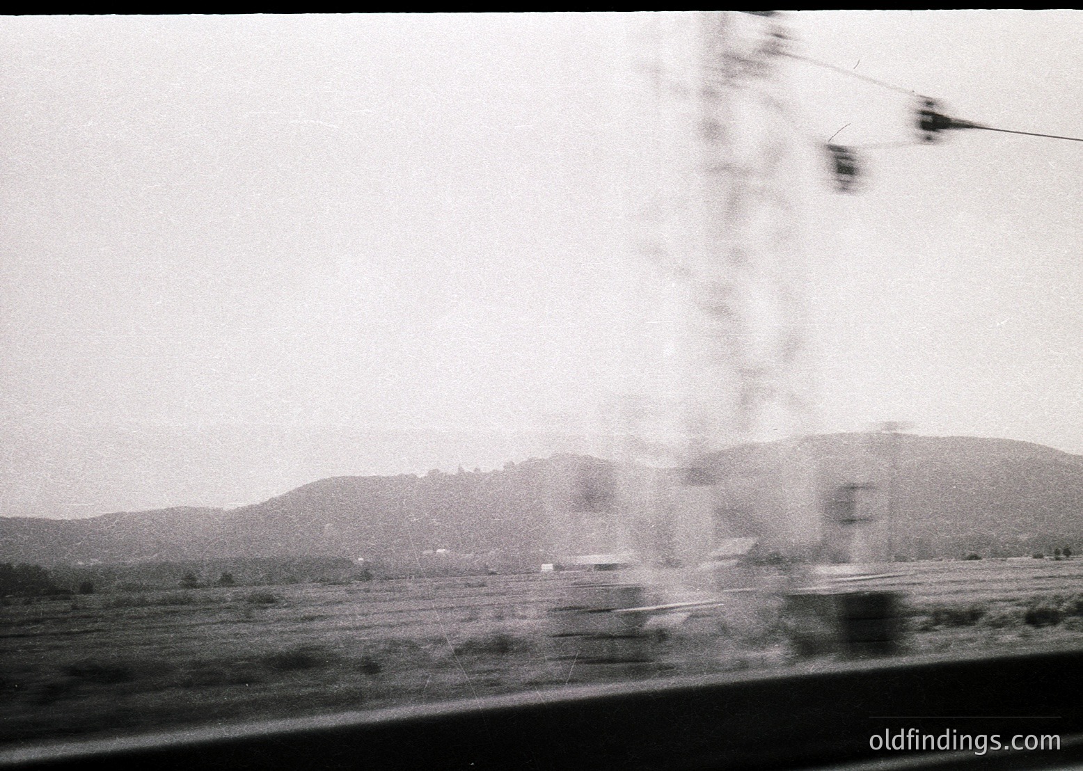Blurred motion shot from a moving vehicle, capturing rural countryside with distant hills and sparse trees. Overhead wires and a blurred bus suggest mid-20th century transportation. Grainy black-and-white film enhances nostalgic, candid atmosphere.