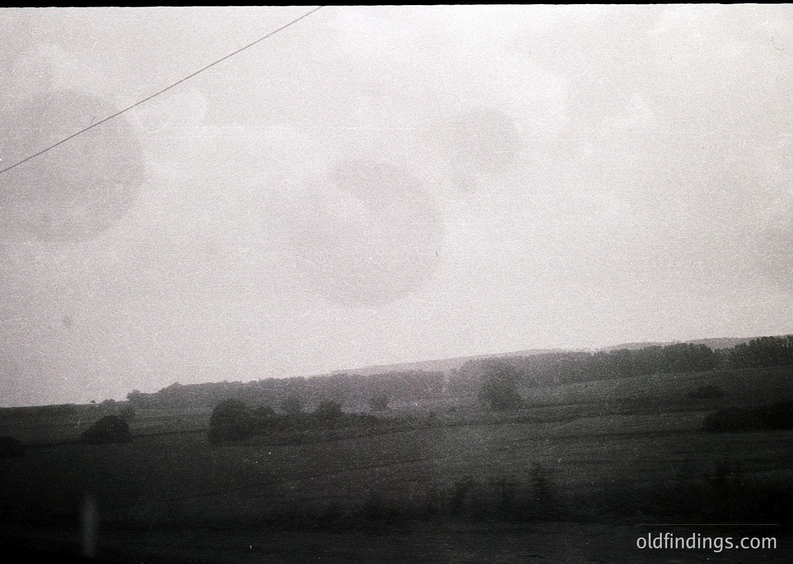 Vintage black-and-white landscape featuring low-lying mist over a flat, open field with sparse trees. Overcast sky and faint power line suggest rural setting. Likely mid-20th century due to monochrome and composition.