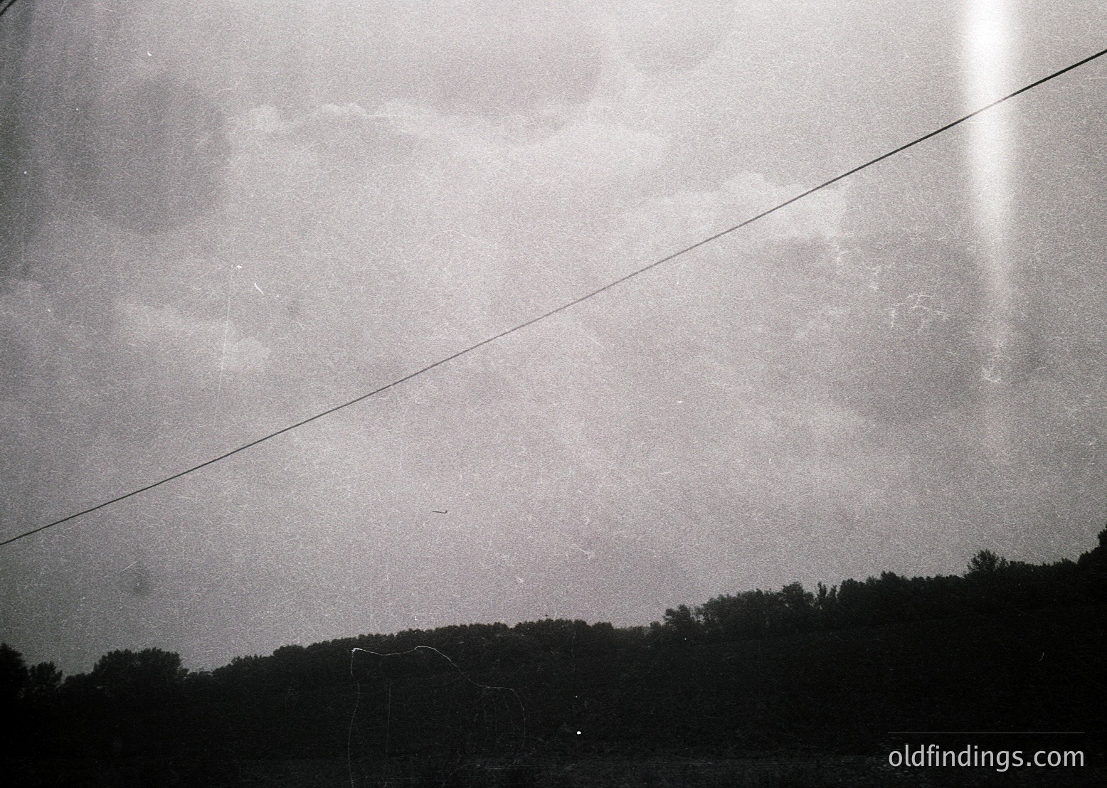 Black-and-white shot of a low-angle sky framed by power lines, evoking mid-20th century documentary style. Dense, overcast clouds obscure horizon, with sparse trees below. Grainy texture suggests vintage film or early digital capture.