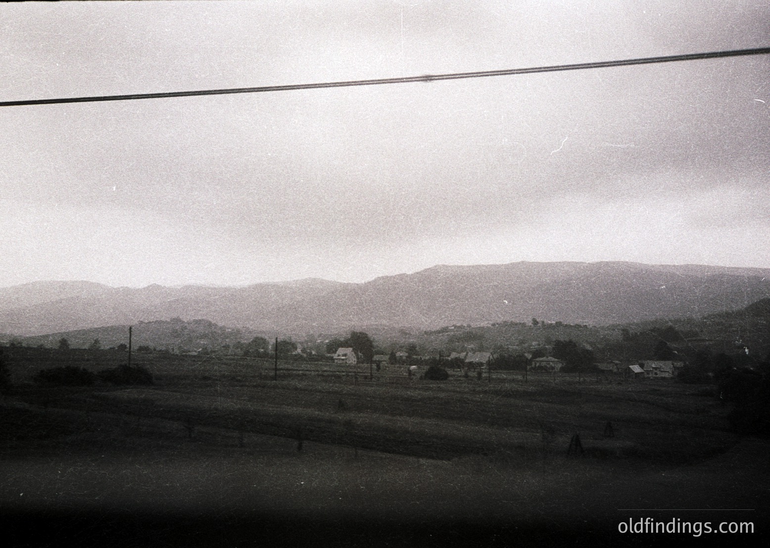Black-and-white rural landscape featuring scattered farmhouses and dense forest on rolling hills. Overcast sky and power line frame the scene, suggesting mid-20th century agricultural life.