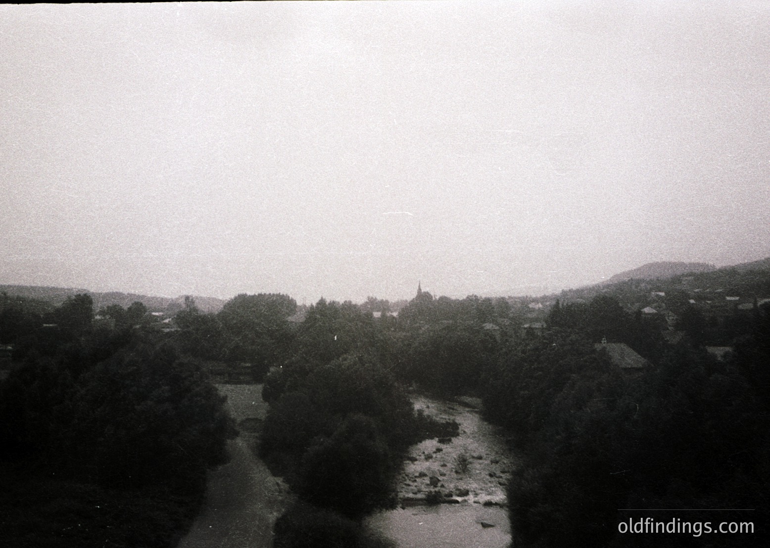 Vintage black-and-white landscape featuring a winding river flanked by dense forest. Distant village with clustered buildings and a prominent church spire on a hillside. Overcast skies enhance atmospheric mood. Likely Eastern European countryside, mid-20th century.
