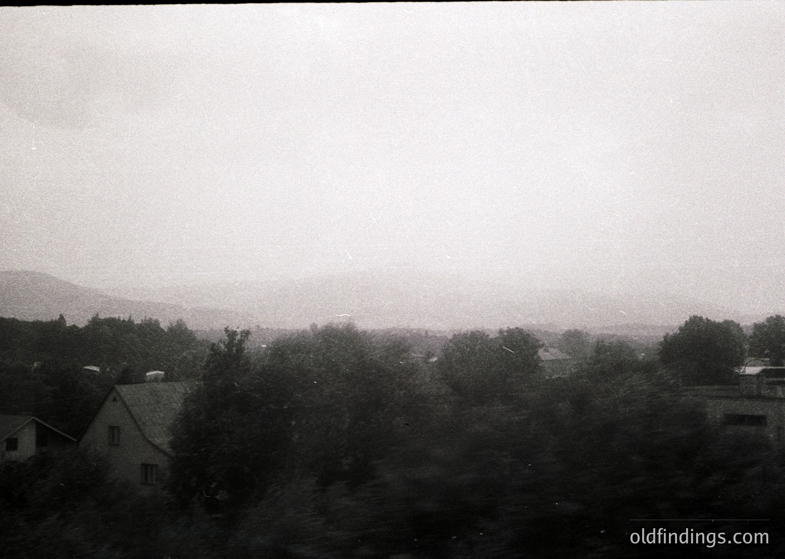 Vague black-and-white landscape shot featuring low-contrast rural scenery. Dense forest in foreground with scattered buildings, likely residential. Distant hills and misty horizon suggest overcast conditions. Style resembles mid-20th century photography.