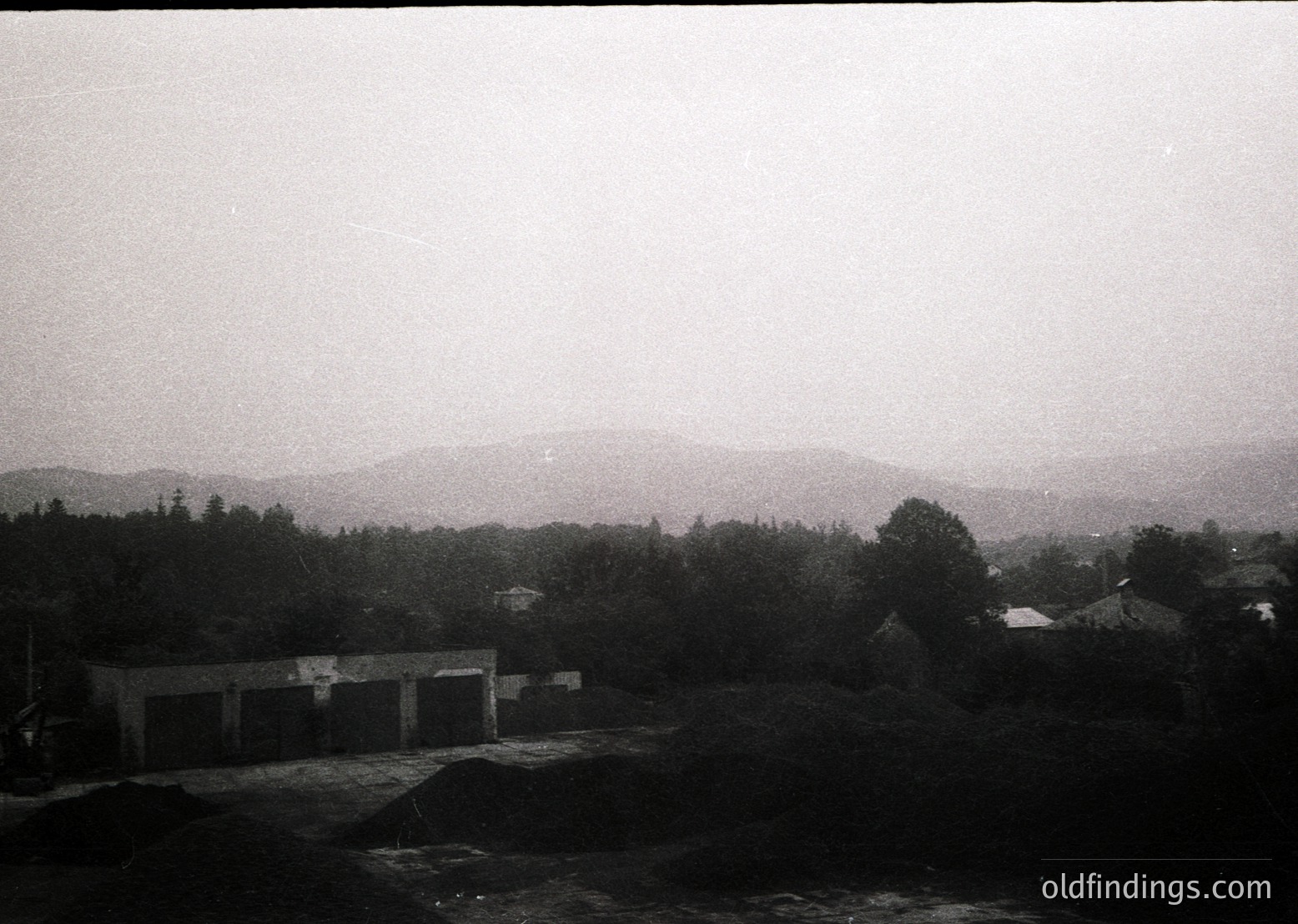 Vintage black-and-white view of a rural landscape featuring a low-lying industrial building complex with flat roofs and minimal windows, likely a factory or warehouse. Dense forest and rolling hills dominate the background under an overcast sky. Mid-20th century architectural style suggests post-war European setting.