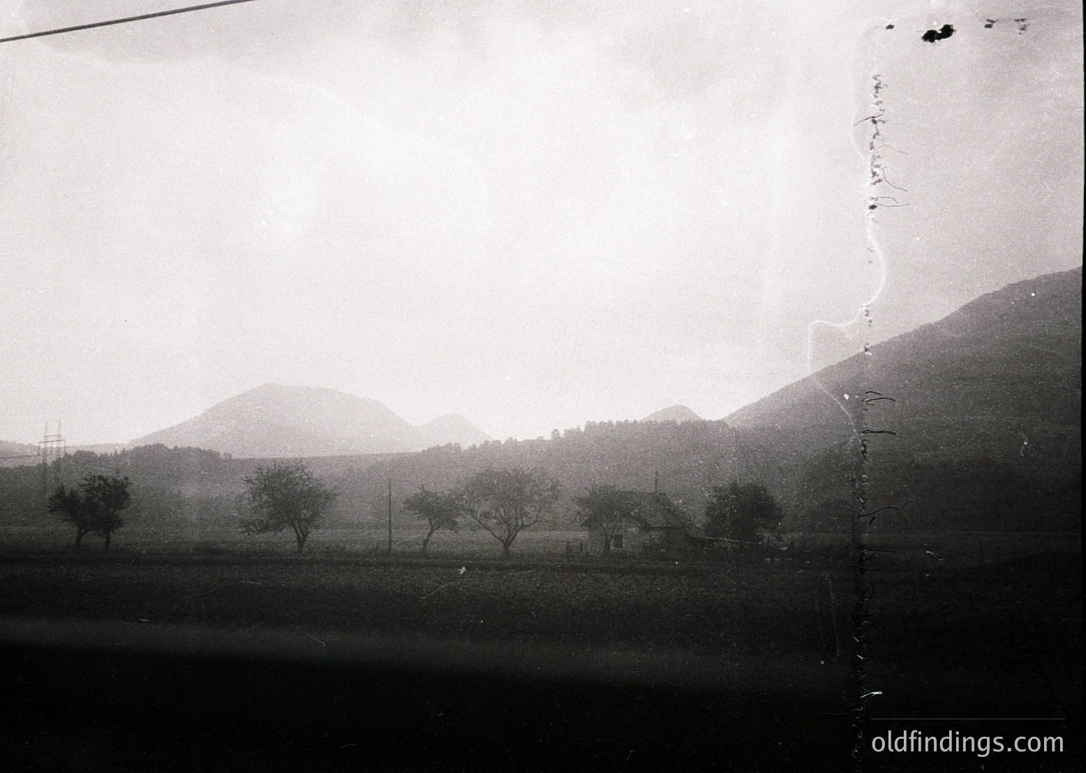 Vintage black-and-white landscape showing sparse trees and rolling hills under overcast skies. Visible power lines suggest rural electrification, likely mid-20th century. Damaged edges indicate aged archival print.