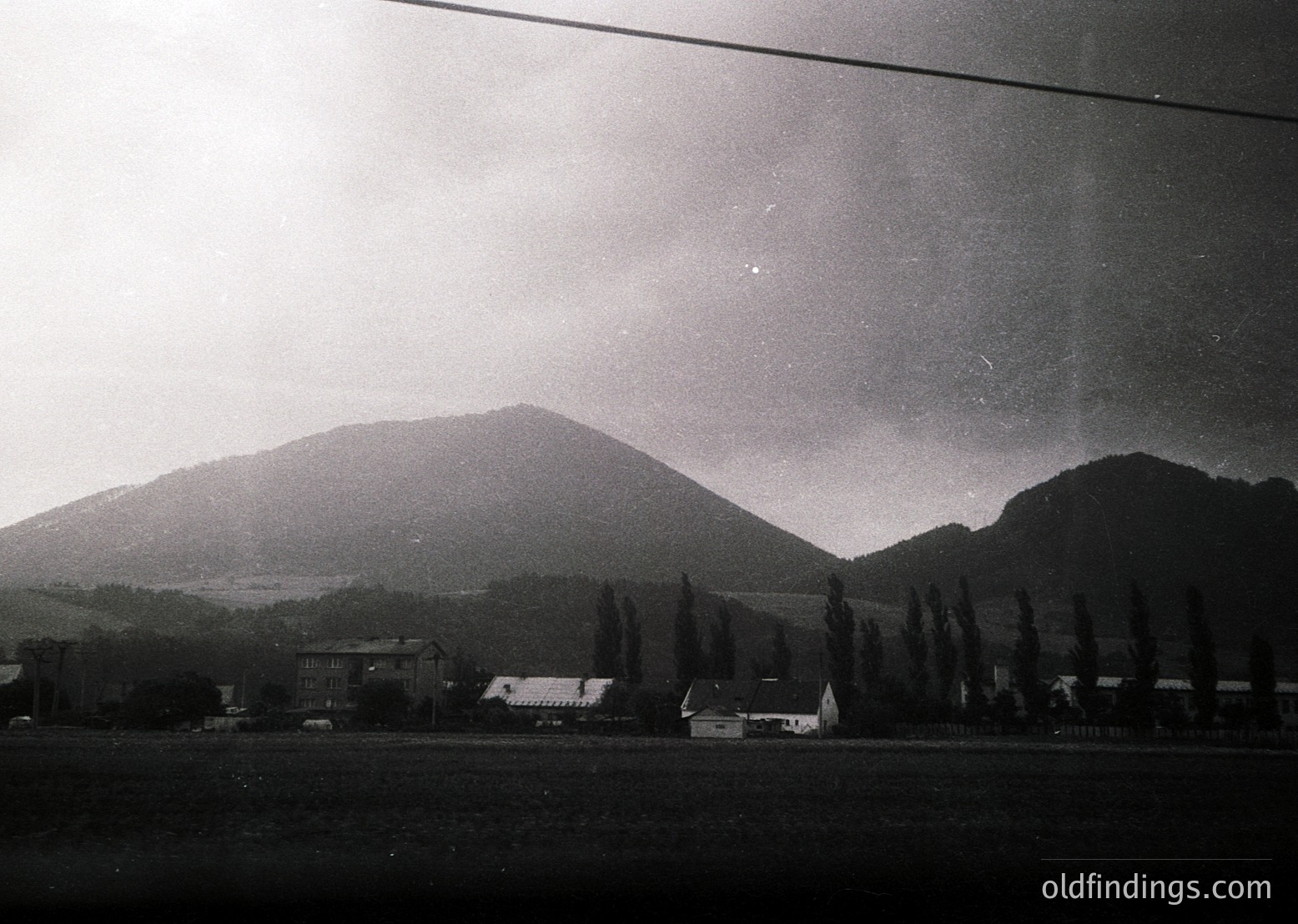 Vintage black-and-white rural landscape featuring rolling hills and scattered farmhouses. Low-angle shot captures misty horizon with dense forest line. Mid-20th century architectural style evident in buildings.