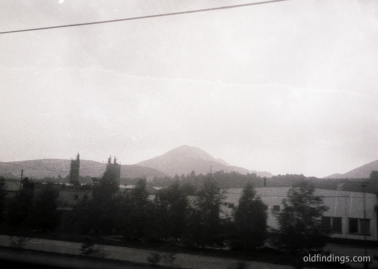Black-and-white train window view of rural landscape with misty mountains in background. Low-lying buildings and dense greenery frame the scene, suggesting a misty, overcast day. Likely Eastern European countryside, 20th century.