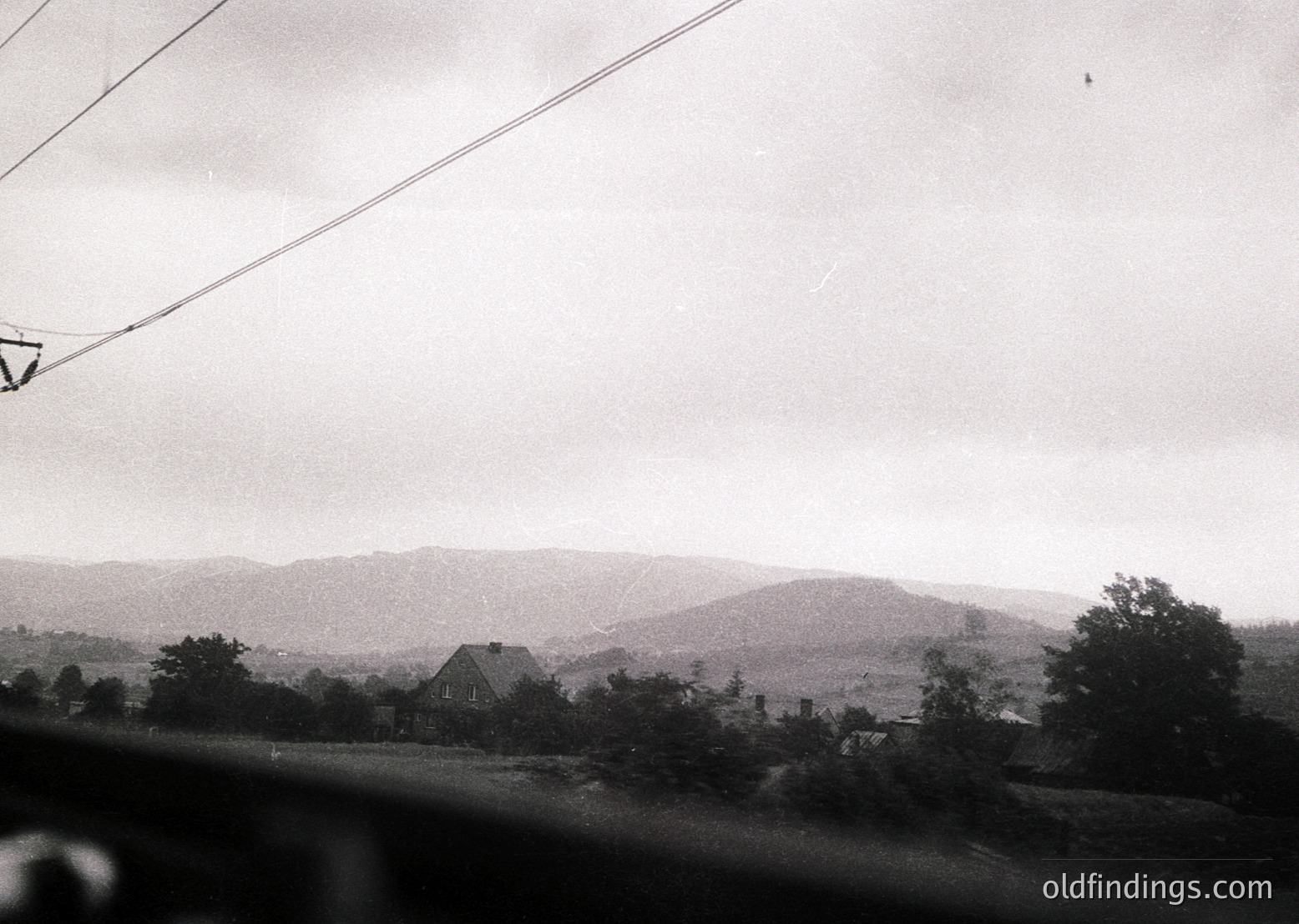 Vintage black-and-white landscape shot of rolling hills and a single-story house framed by trees. Overhead power lines and a blurred foreground suggest motion from a moving vehicle. Rural setting with muted, atmospheric lighting. Likely mid-20th century.