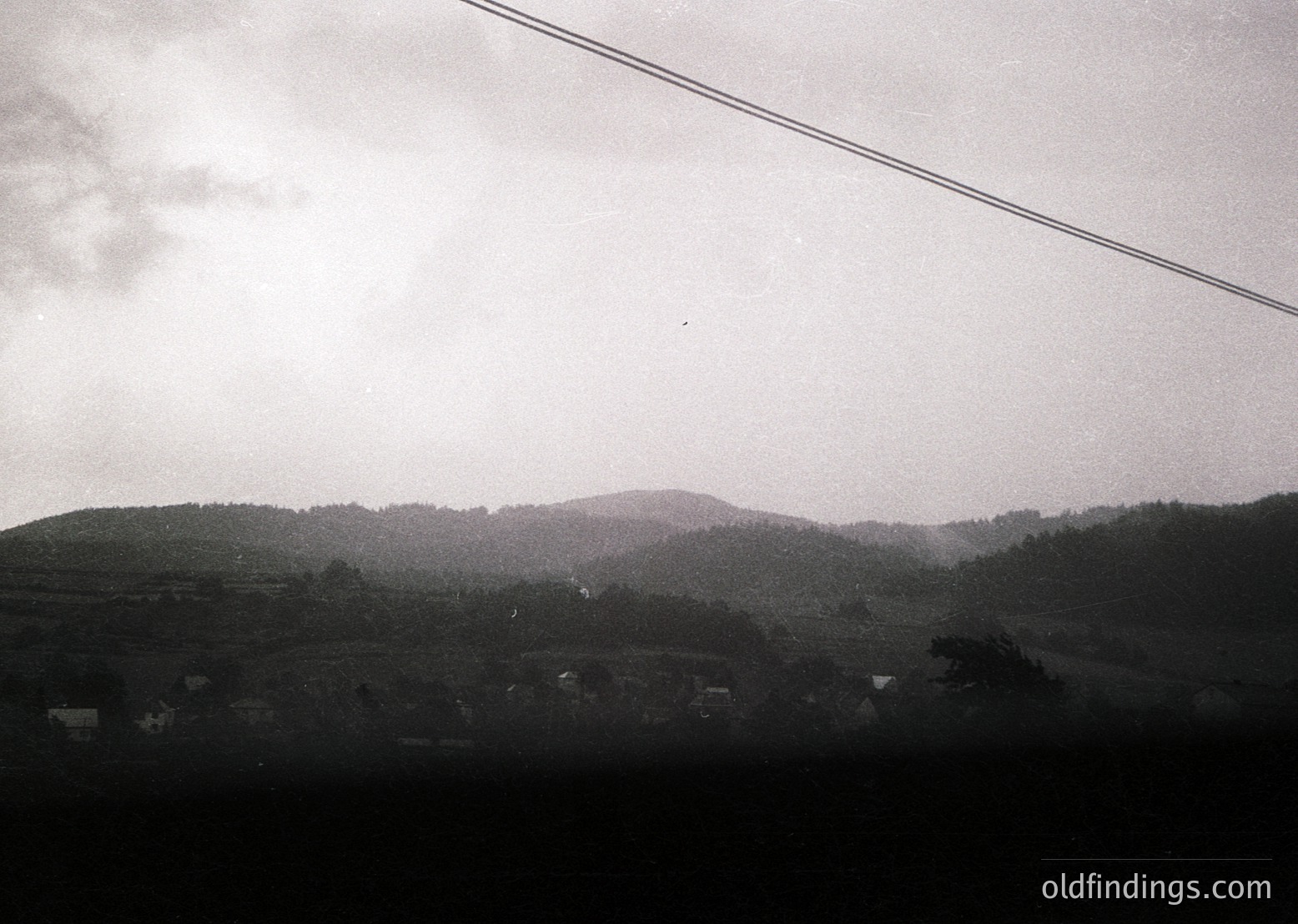 Black-and-white rural landscape featuring rolling hills, scattered farmhouses, and dense forest. Overhead power lines frame the top edge. Likely Eastern European countryside, mid-20th century.