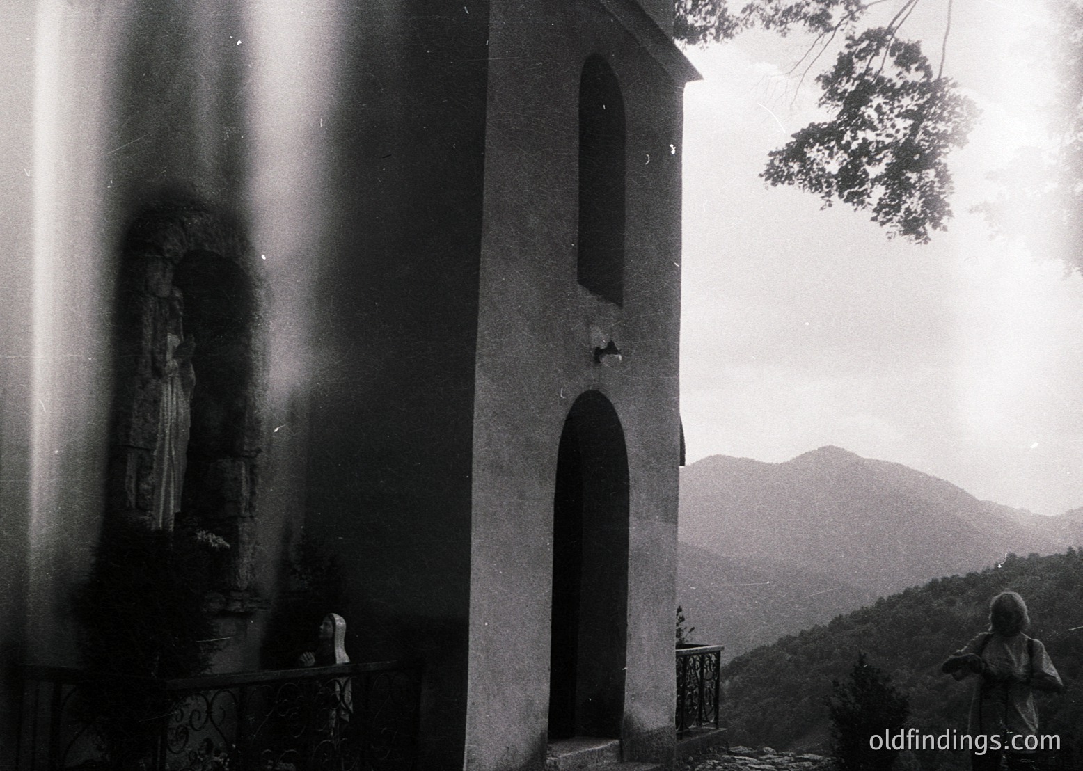 Black-and-white shot of a modest stone chapel with arched doorway, set against misty mountain backdrop. Icon of Virgin Mary inside niche, flanked by ornate wrought-iron railing. Child in traditional dress kneels at altar, suggesting rural Eastern European setting. Likely late 20th century.